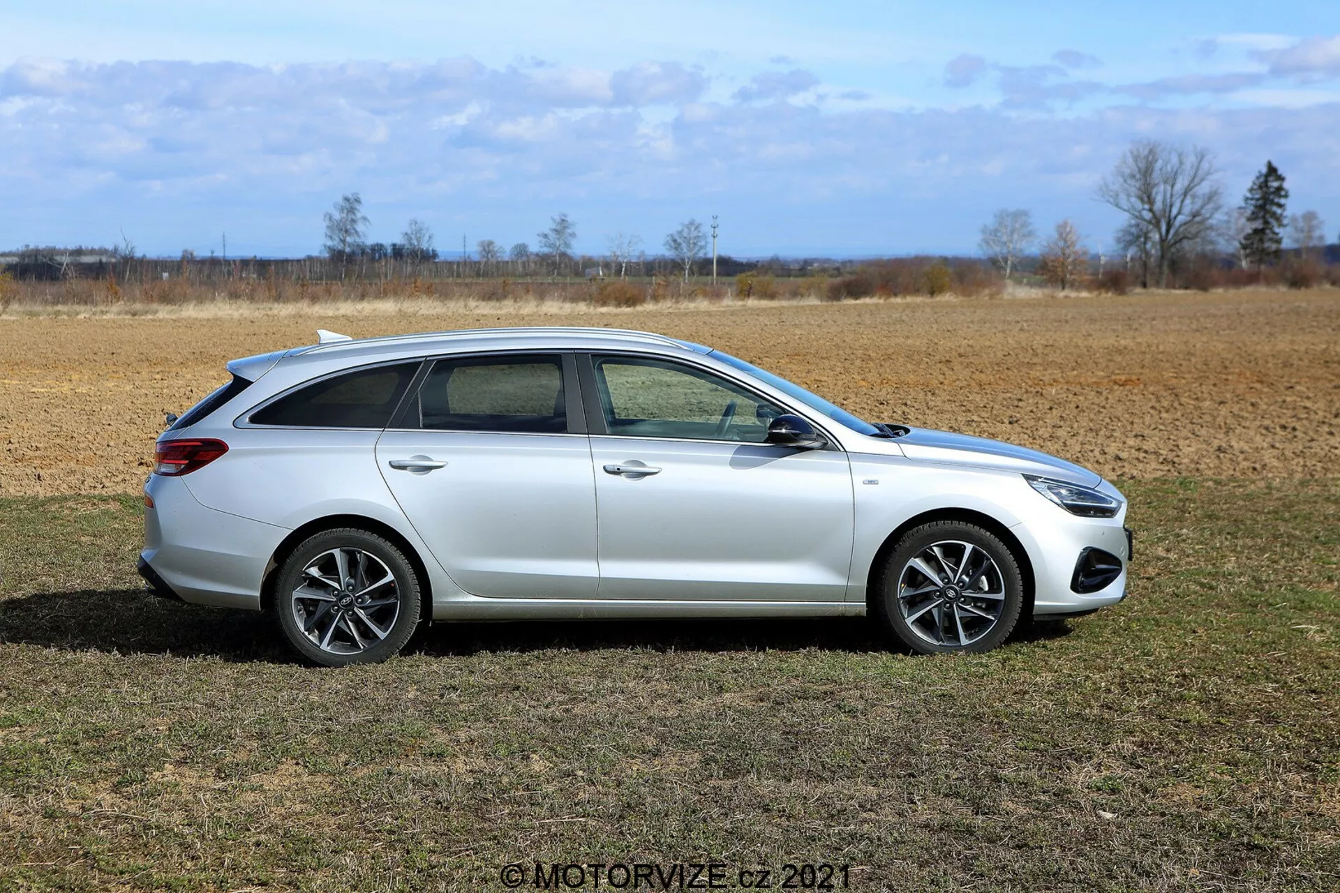 Profil boczny Hyundaia i30 kombi z 2020 roku w błyszczącej białej farbie, zaparkowany na suchym polu trawiastym z jałowym krajobrazem i czystym niebem w tle. Pojazd charakteryzuje się wydłużoną tylną częścią zwiększającą przestrzeń ładunkową, czterema drzwiami, wieloszprychowymi felgami aluminiowymi, dużymi szybami, lusterkami bocznymi, chromowanymi lub metalicznymi wykończeniami oraz zintegrowanymi relingami dachowymi.