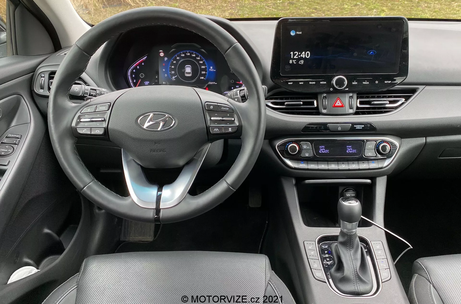 Widok wnętrza Hyundai i30 Kombi 2020 z fotela kierowcy, ukazujący nowoczesny kokpit z kierownicą ozdobioną logo Hyundai z przyciskami sterującymi funkcjami takimi jak tempomat, audio i połączenia bezprzewodowe. Cyfrowy wyświetlacz deski rozdzielczej pokazuje prędkość i inne informacje o pojeździe. Na środku deski rozdzielczej znajduje się duży ekran dotykowy systemu infotainment do nawigacji i mediów, obok fizycznych przycisków klimatyzacji, co sugeruje dwu-strefowy system klimatyzacji. Konsola środkowa zawiera dźwignię zmiany biegów z skórzanym mieszkiem i portem USB, co podkreśla zaawansowane funkcje łączności i technologii.