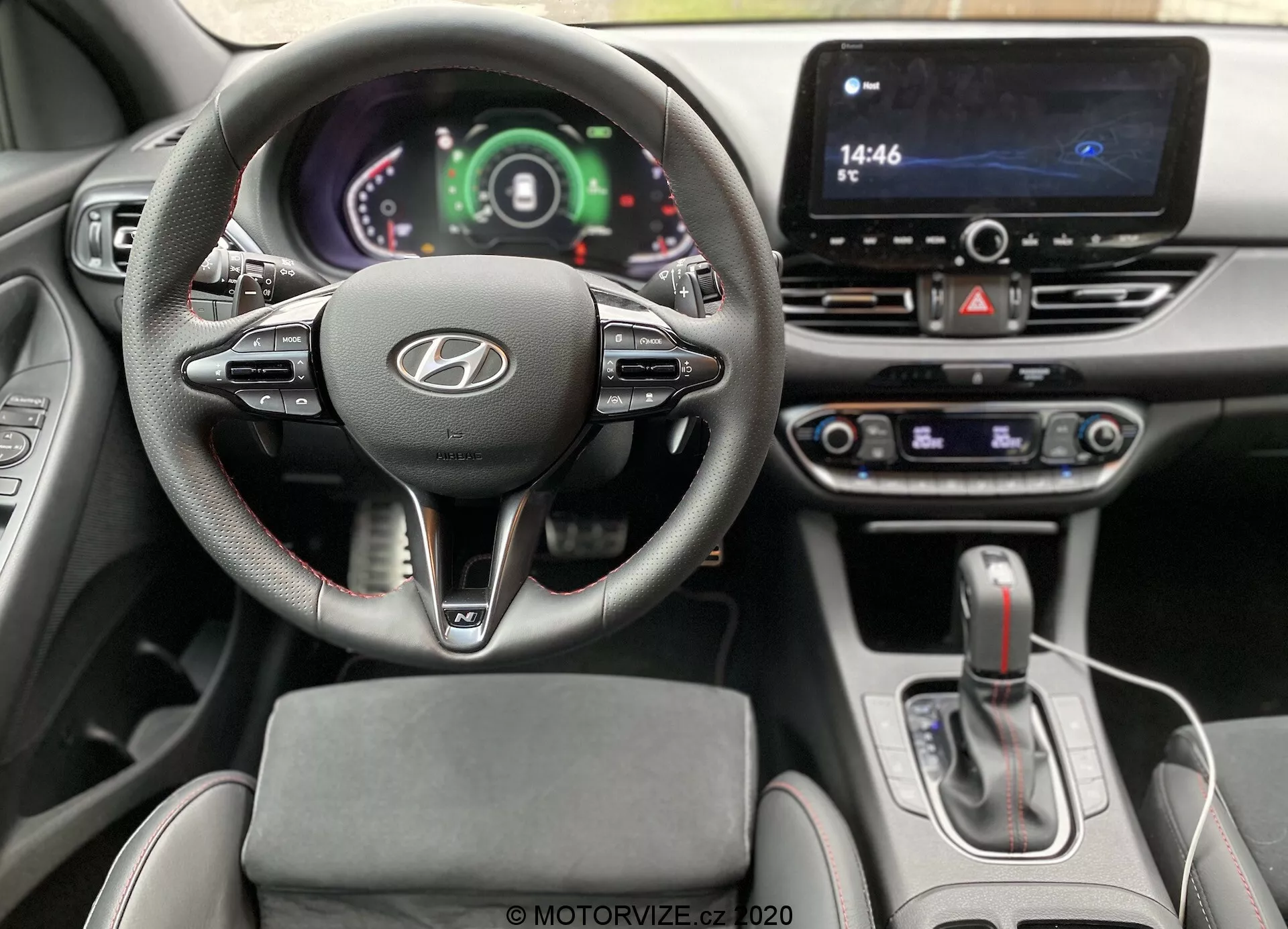 Wnętrze z perspektywy kierowcy wewnątrz Hyundai i30 2020 (i30-kombi), przedstawiające kierownicę z logo Hyundai, zamontowane kontrolki i cyfrowy zestaw wskaźników. Widoczne są system infotainment z ekranem dotykowym, selektor automatycznej skrzyni biegów i pokrętła sterowania klimatyzacją, wskazujące na zaawansowaną technologię i nowoczesny design.