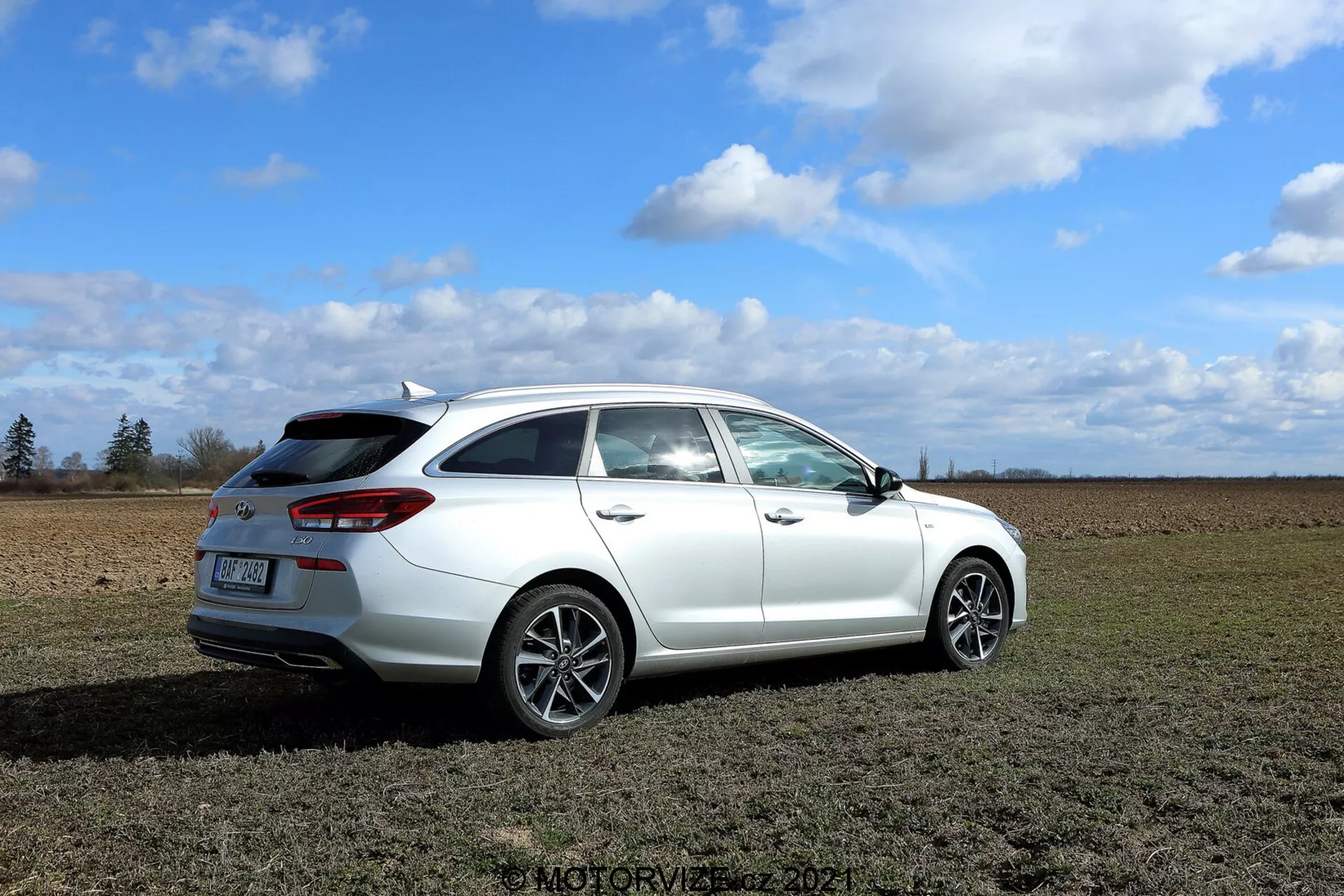 Szczegółowy widok białego Hyundai i30 kombi z 2020 roku, zaparkowanego na trawiastym polu z częściowo pochmurnym niebem w tle. Zdjęcie zrobione pod kątem trzech czwartych z tyłu, ukazujące takie elementy wzornictwa jak nowoczesne tylne światła obejmujące boki pojazdu, tylna szyba z wycieraczką, mały spoiler dachowy, wieloszprychowe felgi aluminiowe oraz lusterko boczne z możliwymi nowoczesnymi funkcjami. Widoczne są linie zamknięcia tylnej drzwi dla pasażerów i klapy bagażnika, sugerujące co najmniej cztery drzwi. Chociaż wydech nie jest wyraźnie widoczny, wydaje się być dyskretny. Samochód jest wyraźnie ostro ustawiony, podkreślając jego design i funkcje, z otaczającym środowiskiem tworzącym kontekstualną scenerię.