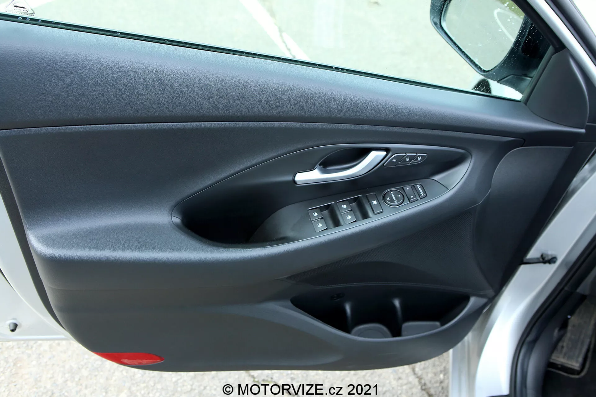 Panel drzwi wewnętrznych Hyundai i30 2020 (i30-kombi) widziany pod kątem skośnym, ukazujący srebrną chromowaną klamkę, ergonomiczne przyciski sterowania szybami elektrycznymi, elektroniczny przycisk blokady drzwi, kieszeń na przechowywanie oraz dwuteksturowane wykończenie drzwi z miękkim górnym panelem i sztywną dolną częścią z tworzywa sztucznego w ciemnych kolorach.