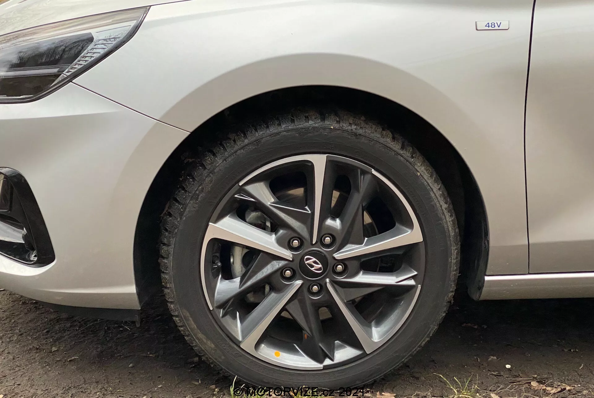 Zbliżenie przedniego koła Hyundai i30 Kombi 2020, ukazujące nowoczesną felgę aluminiową z wieloszprychowym wzorem i widocznymi tarczami hamulcowymi za szprychami. Ujęcie, wykonane z niskiego kąta blisko ziemi, obejmuje część bocznej karoserii w metalicznym, srebrnym kolorze, z widocznym emblematem 4WD wskazującym na napęd na cztery koła. Obraz podkreśla czysty design samochodu z zakrzywionymi liniami i drobnymi charakterystycznymi detalami nad nadkolem.