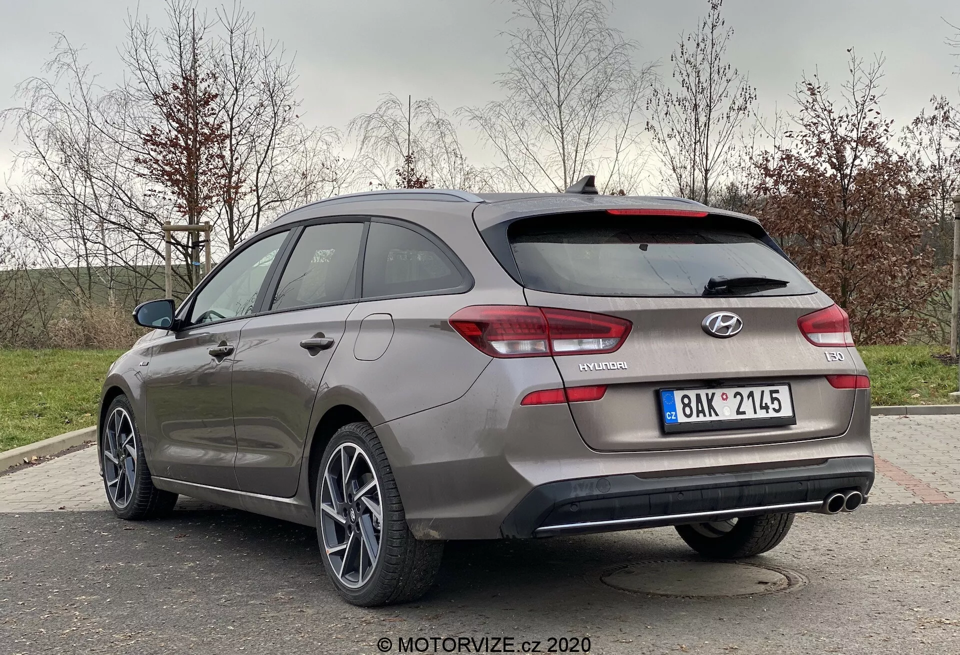 Obraz szarego Hyundai i30 2020 (i30-kombi) z perspektywy trzech czwartych tyłu. Samochód zaparkowany na świeżym powietrzu na przedmieściach lub w parku, ukazując tylną i boczną stronę projektową. Widoczne elementy to hatchback z tylnym spojlerem, tylne światła, odznaka 'i30', odznaka 'N Line', podwójne rury wydechowe i wieloszprychowe felgi aluminiowe. Otoczenie sugeruje trawiastą okolicę z drzewami w tle.