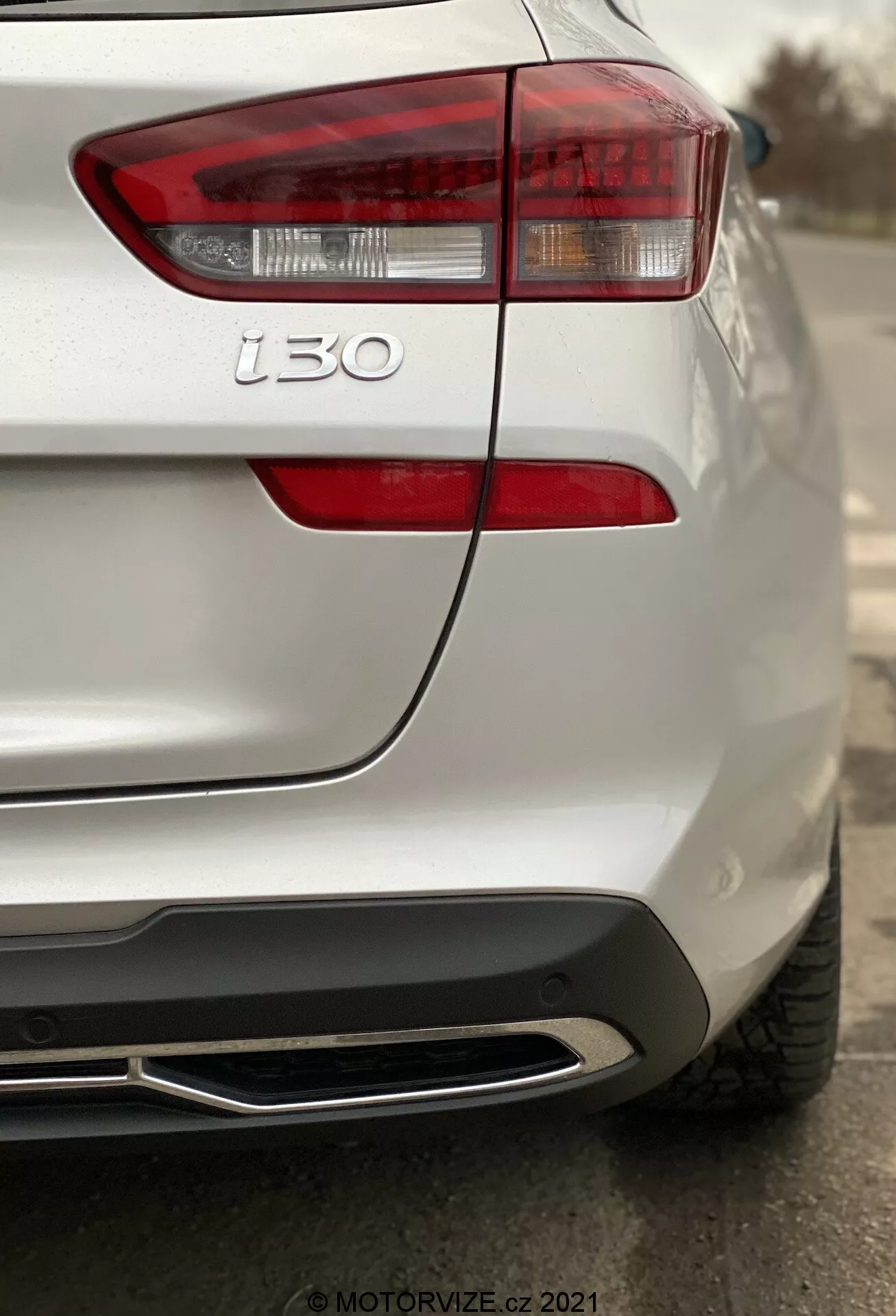 Zbliżony widok tylnej części białego Hyundai i30 2020 (Kombi) skoncentrowany na lewym zespole tylnych świateł i lewej tylnej kwaterze, ukazujący nowoczesne linie projektowe, przejrzyste soczewki świateł hamowania, kierunkowskazy, odznakę i30, szczegółową strukturę lakieru, czujnik parkowania, płaską integrację i chromowaną końcówkę wydechu, z płytką głębią ostrości podkreślającą tylny design samochodu.