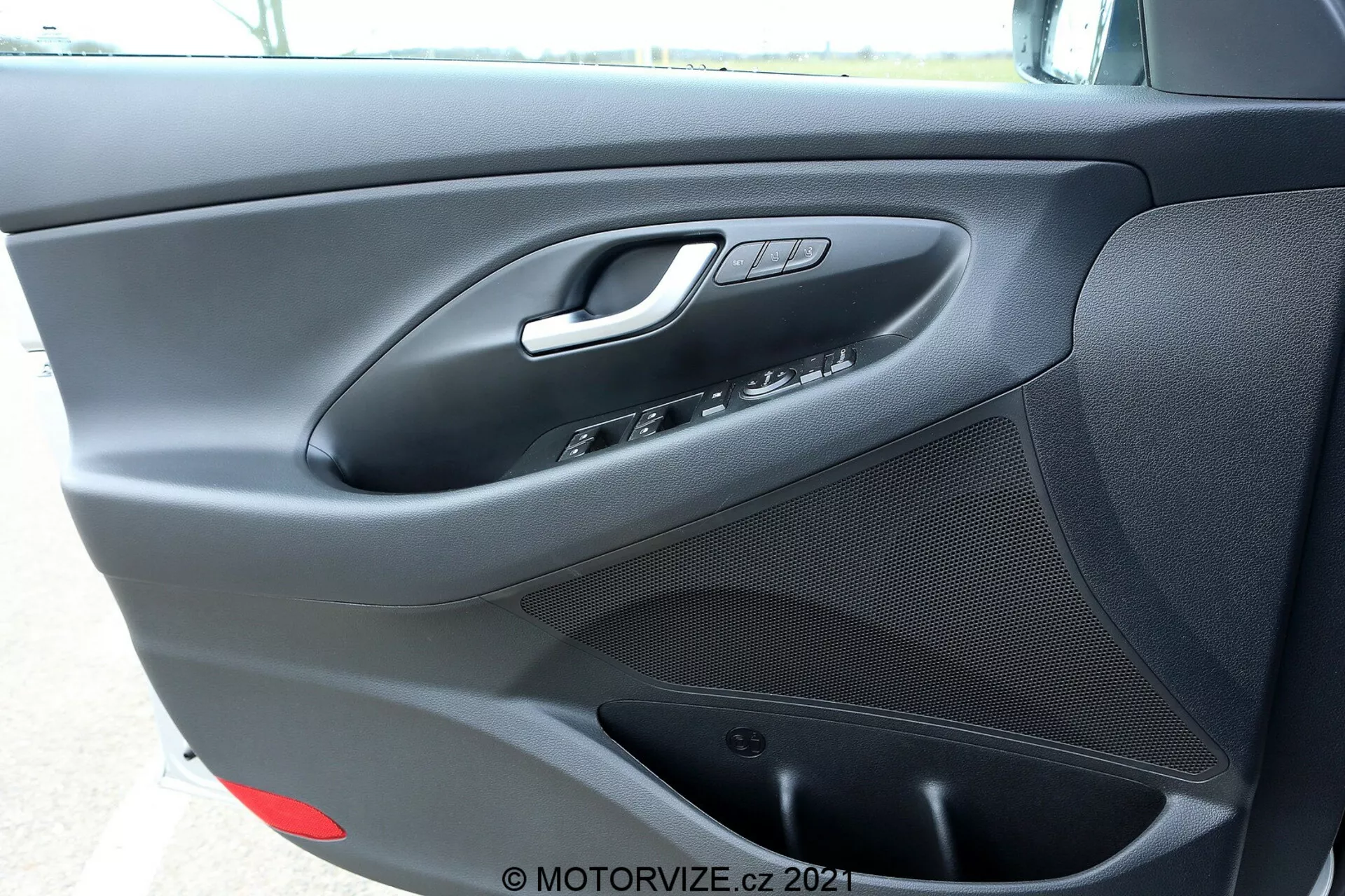 Zbliżenie na wewnętrzny panel drzwi po stronie kierowcy Hyundai i30 Kombi 2020, pokazujący srebrny uchwyt drzwiowy, przyciski sterowania oknami i lusterkami, przycisk blokady drzwi, zintegrowany głośnik na dole oraz teksturowane, matowe czarne wykończenie z prawdopodobnie małym schowkiem na dole.