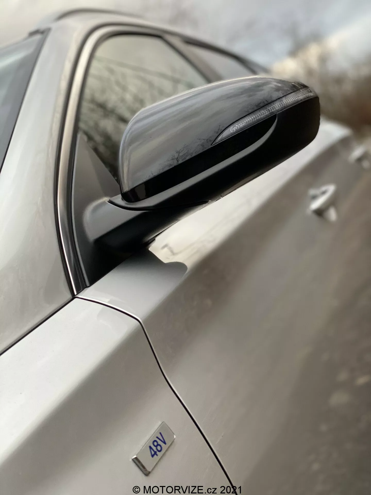 Kątowy zbliżony obraz bocznego lusterka Hyundai i30 2020, wykonany od tylnej części samochodu w kierunku przodu. Ujęcie koncentruje się na zespole lusterek, w tym nowoczesnej obudowie z wbudowanymi kierunkowskazami i powierzchnią odblaskową. Drzwi kierowcy i jasny kolor nadwozia auta są rozmyte, co podkreśla design i szczegóły lusterka. Widoczna jest odznaka '4DV', ale jej konkretne znaczenie nie jest jasne.