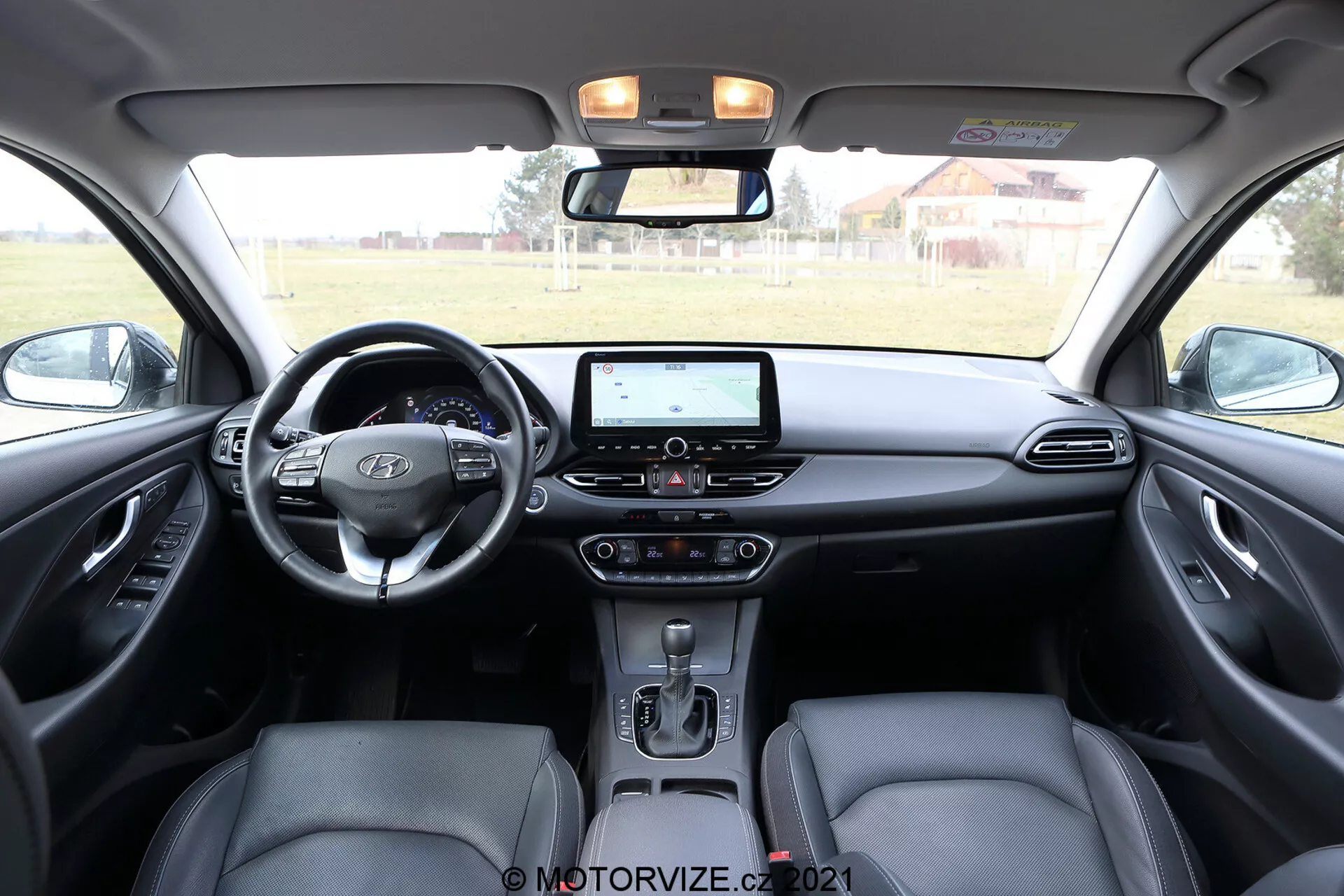 Wnętrze Hyundai i30 2020 Kombi przedstawiające obszar przedniej deski rozdzielczej, widziane z tylnego siedzenia. Zdjęcie pokazuje nowoczesną, wielofunkcyjną kierownicę z przyciskami sterującymi, dotykowy system infotainment z nawigacją i opcjami multimedialnymi, klimatyzację dwustrefową, automatyczną dźwignię zmiany biegów oraz ergonomiczne przednie siedzenia z zagłówkami. Dwukolorowy wystrój wnętrza kontrastuje ciemne i jasne odcienie, dodając głębi. Panele drzwi odzwierciedlają design z możliwymi kratkami głośników i przyciskami sterującymi do elektrycznych szyb i lusterek. Konsola górna zawiera oświetlenie kabiny i przyciski do dodatkowych funkcji.