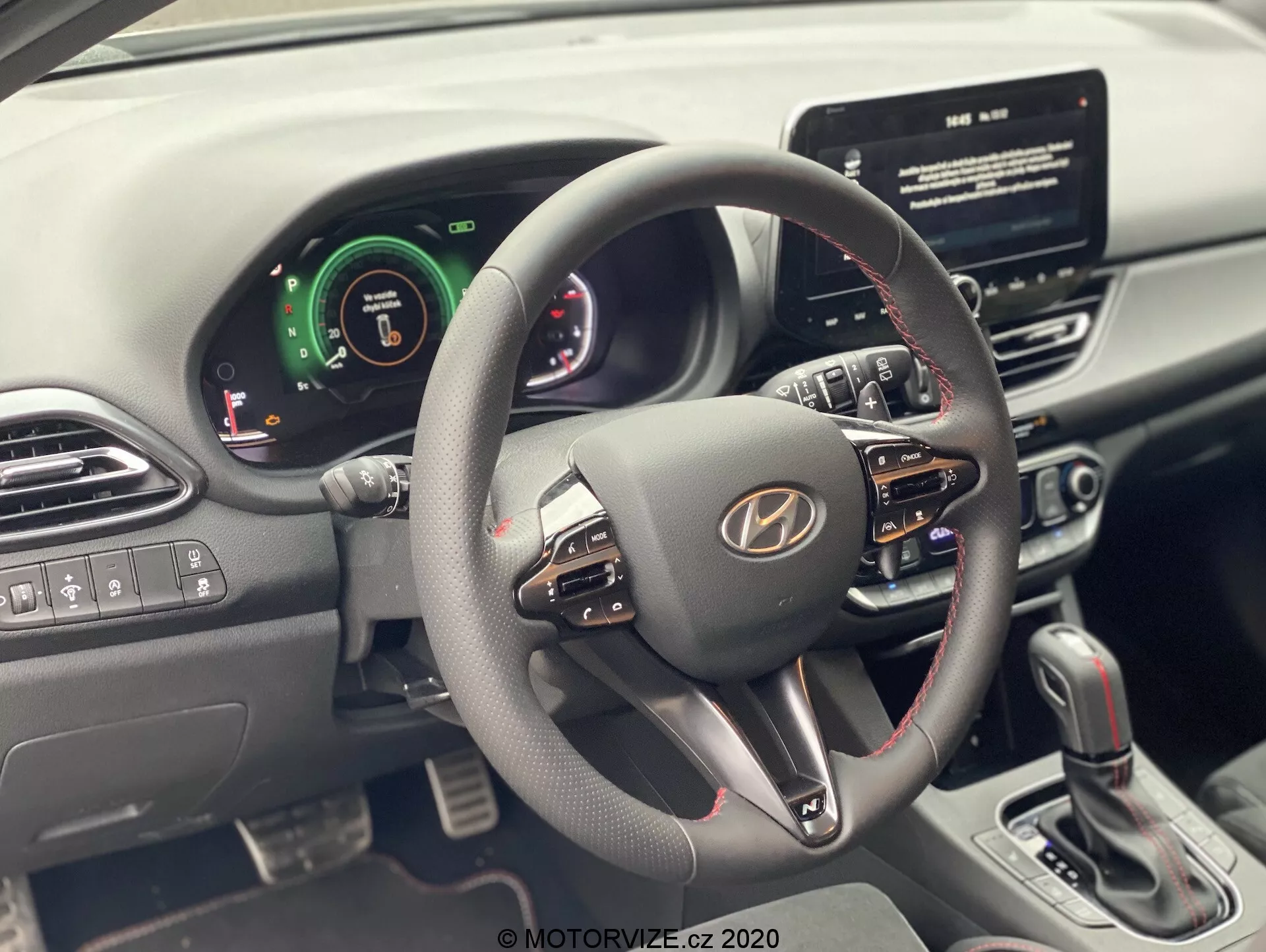 Widok wnętrza Hyundai i30 2020 (i30-kombi) z perspektywy siedzenia kierowcy, koncentrujący się na kierownicy i desce rozdzielczej. Nowoczesna kierownica ma wiele przycisków sterujących do regulacji głośności, komendy głosowe i rozmowy zestawu głośnomówiącego. Zestaw wskaźników łączy elementy cyfrowe i analogowe, wskazując na zaawansowane systemy wspomagania kierowcy. Widoczny jest centralny ekran dotykowy systemu infotainment, sugerujący nawigację i odtwarzanie multimediów. Konsola centralna pokazuje przyciski sterowania klimatyzacją i dźwignię zmiany biegów automatycznej skrzyni biegów. Kontrola okien i lusterek bocznych znajduje się na drzwiach kierowcy.
