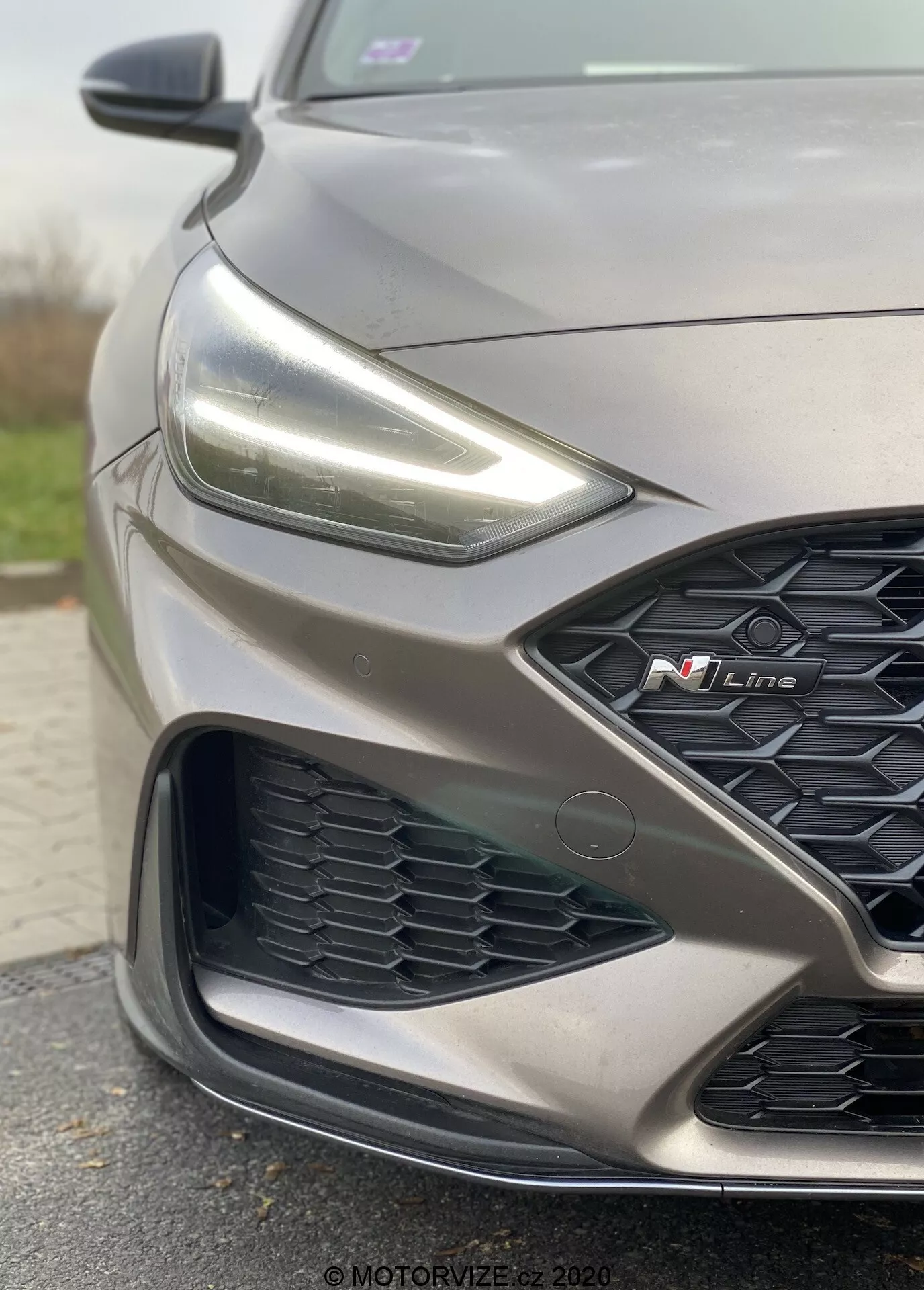 Zbliżenie, ukośny widok skupiający się na przedniej, lewej stronie szarego Hyundai i30 2020 N Line. Obraz podkreśla sportowy design, w tym subtelny kontur maski, ostre reflektory LED, kratkę w kształcie plastra miodu z widocznym czerwonym znakiem 'N Line' oraz skomplikowany zderzak z zaawansowanymi czujnikami sugerującymi nowoczesne systemy wspomagania kierowcy.