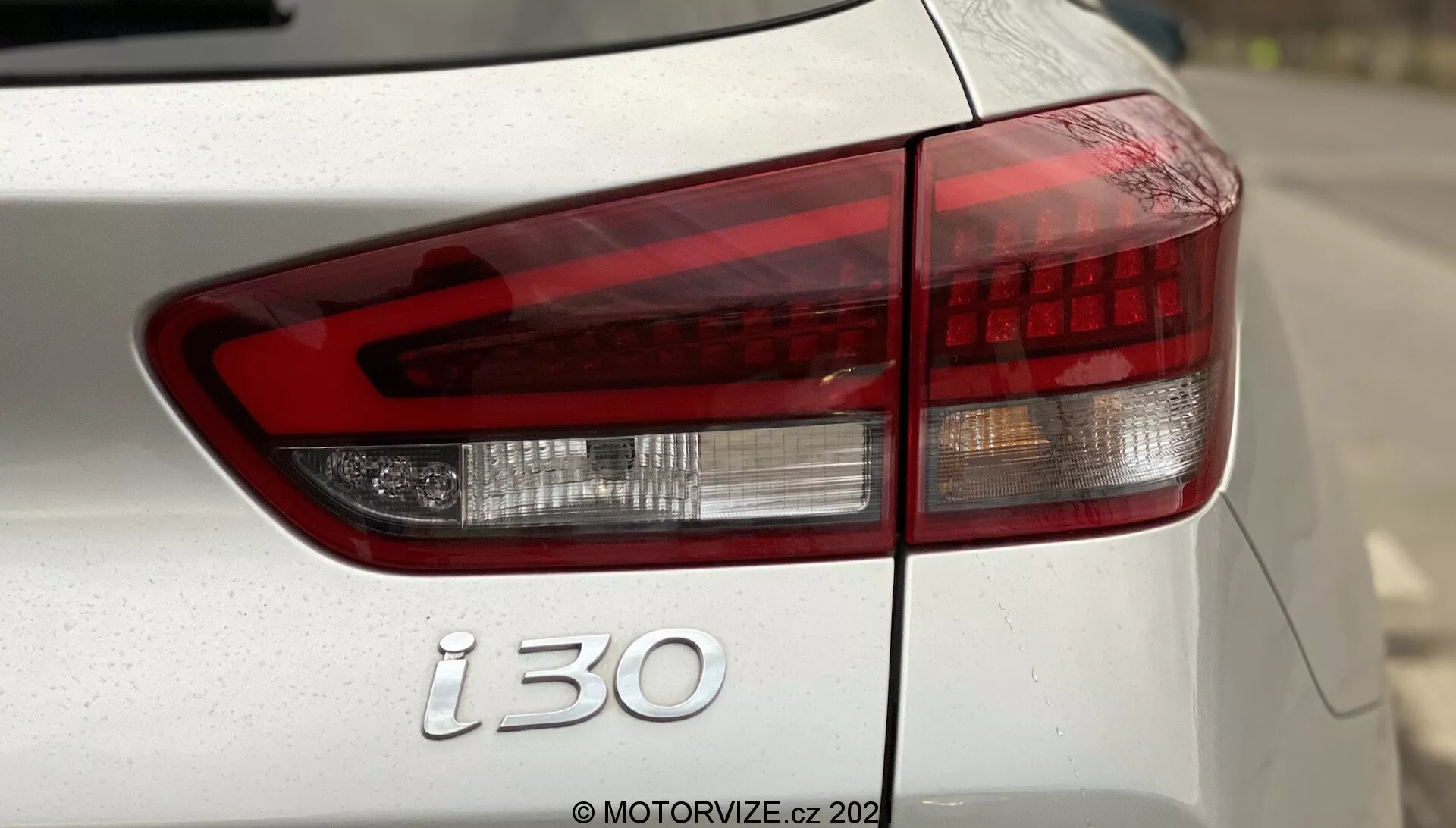 Zbliżenie tylnej lewej strony Hyundai i30 Kombi 2020, ukazujące nowoczesny zestaw świateł tylnych z wyraźnym rozróżnieniem między czerwonym światłem hamowania, białym światłem cofania i pomarańczowym kierunkowskazem zgodnie z europejskimi specyfikacjami. Na zdjęciu widać część klapy bagażnika, tylnego zderzaka oraz emblemat 'i30', z lakierem prawdopodobnie w kolorze srebrnym lub białym. Mała głębia ostrości podkreśla światła tylne i emblemat, z rozmytym tłem oraz widocznym znakiem wodnym 'MOTORVIZE.cz 202'.
