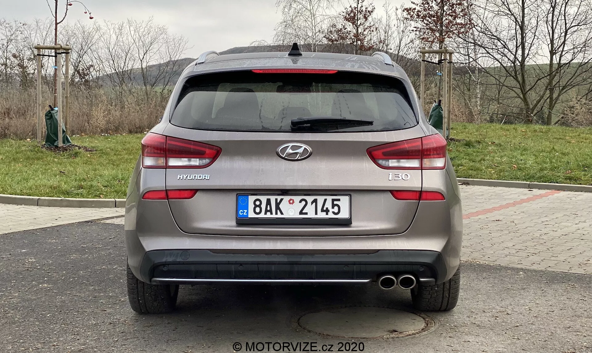 Zdjęcie przedstawia widok z tyłu Hyundai i30 Kombi 2020. Samochód jest zaparkowany, a ujęcie wykonano z bezpośredniego kąta, mniej więcej na wysokości bagażnika pojazdu. Obraz skoncentrowany na pojeździe, daje wyraźny i symetryczny widok tylnej części samochodu, w tym logo Hyundai, nazwę modelu 'i30' oraz oznaczenie wersji 'CRDi', co wskazuje na silnik diesel z bezpośrednim wtryskiem Common Rail. Widoczne szczegóły obejmują europejską tablicę rejestracyjną, poziome światła tylne, tylne czujniki, podwójne końcówki wydechu, tylna wycieraczka, antenę w kształcie płetwy rekina oraz potencjalnie tylna kamera w pobliżu obszaru tablicy rejestracyjnej.