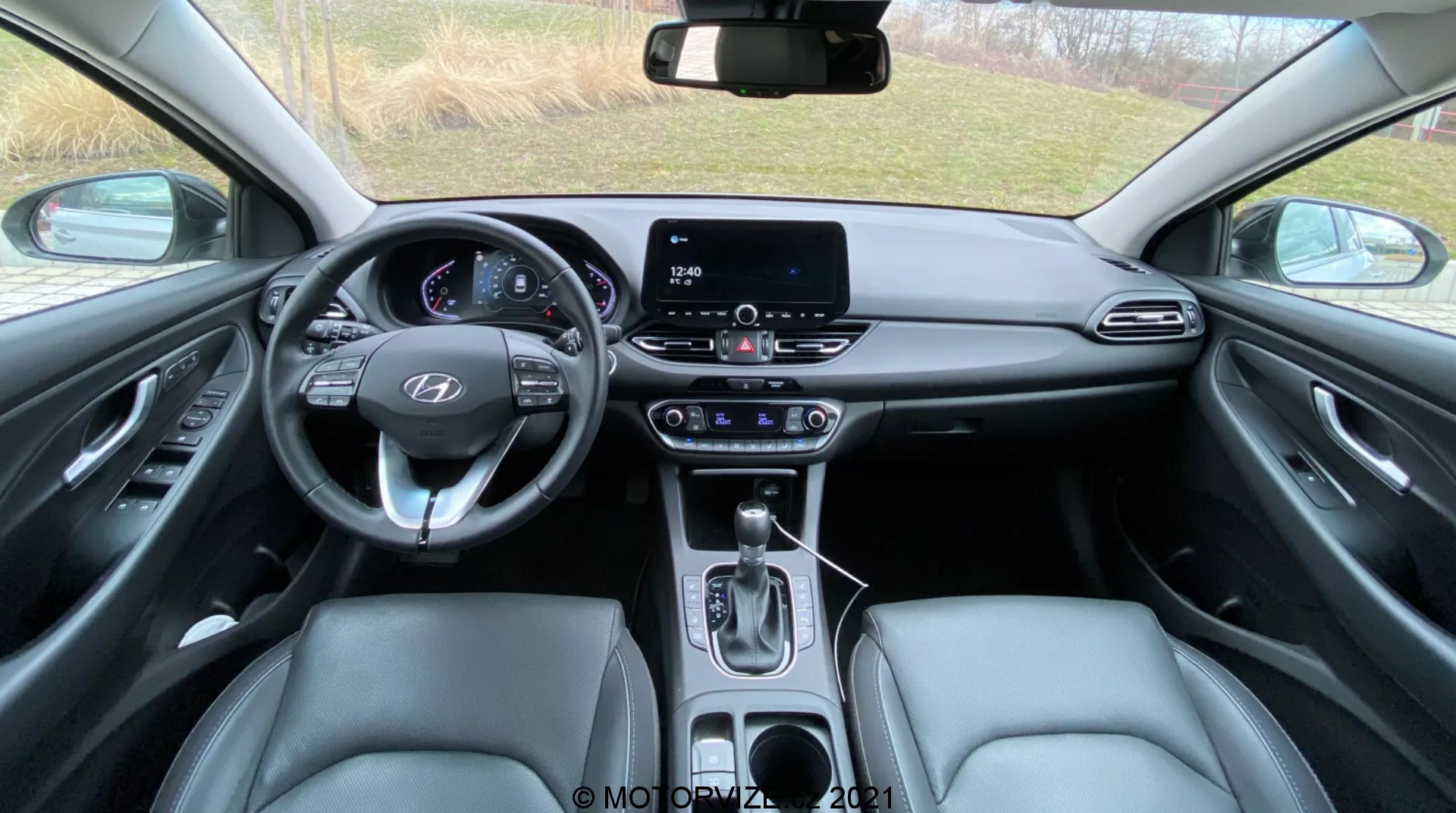 Widok wnętrza Hyundai i30 2020 (i30-Kombi) z tylnego siedzenia, prezentujący wielofunkcyjną kierownicę z zamontowanymi przyciskami, cyfrowy zestaw wskaźników wskazujący tryb parkowania oraz otwarte drzwi kierowcy, dotykowy system informacji i rozrywki, automatyczną dźwignię zmiany biegów, przednie siedzenia wykończone skórą, szczegółowe panele drzwi z elektrycznymi kontrolkami, różnorodne materiały wnętrza obejmujące syntetyczną skórę i metaliczne akcenty, górny panel z miejscem na okulary przeciwsłoneczne i przyciskami, auto-ściemniające lusterko wsteczne z dodatkowymi guzikami oraz częściowo widoczne dywaniki podłogowe.