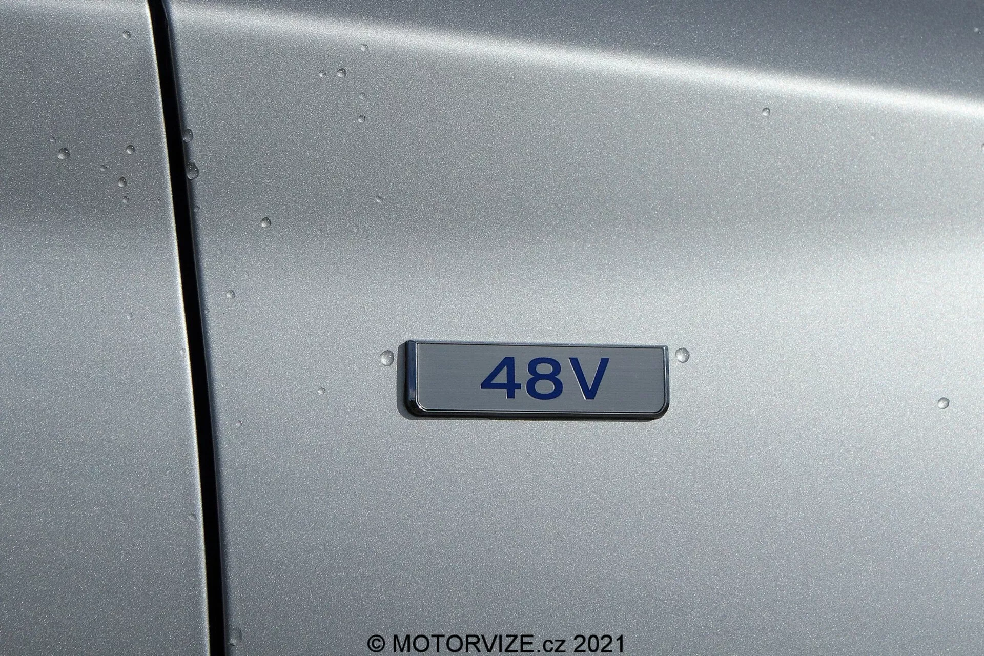 Zbliżenie metalicznie szarego panelu bocznego Hyundai i30 2020 (i30-Kombi) z emblematem '48V', wskazującym na 48-woltowy system mild-hybrid, z powierzchnią pokrytą kroplami deszczu, bez widoku panoramicznego, neutralnym oświetleniem, czarnym tłem i srebrnymi cyframi.