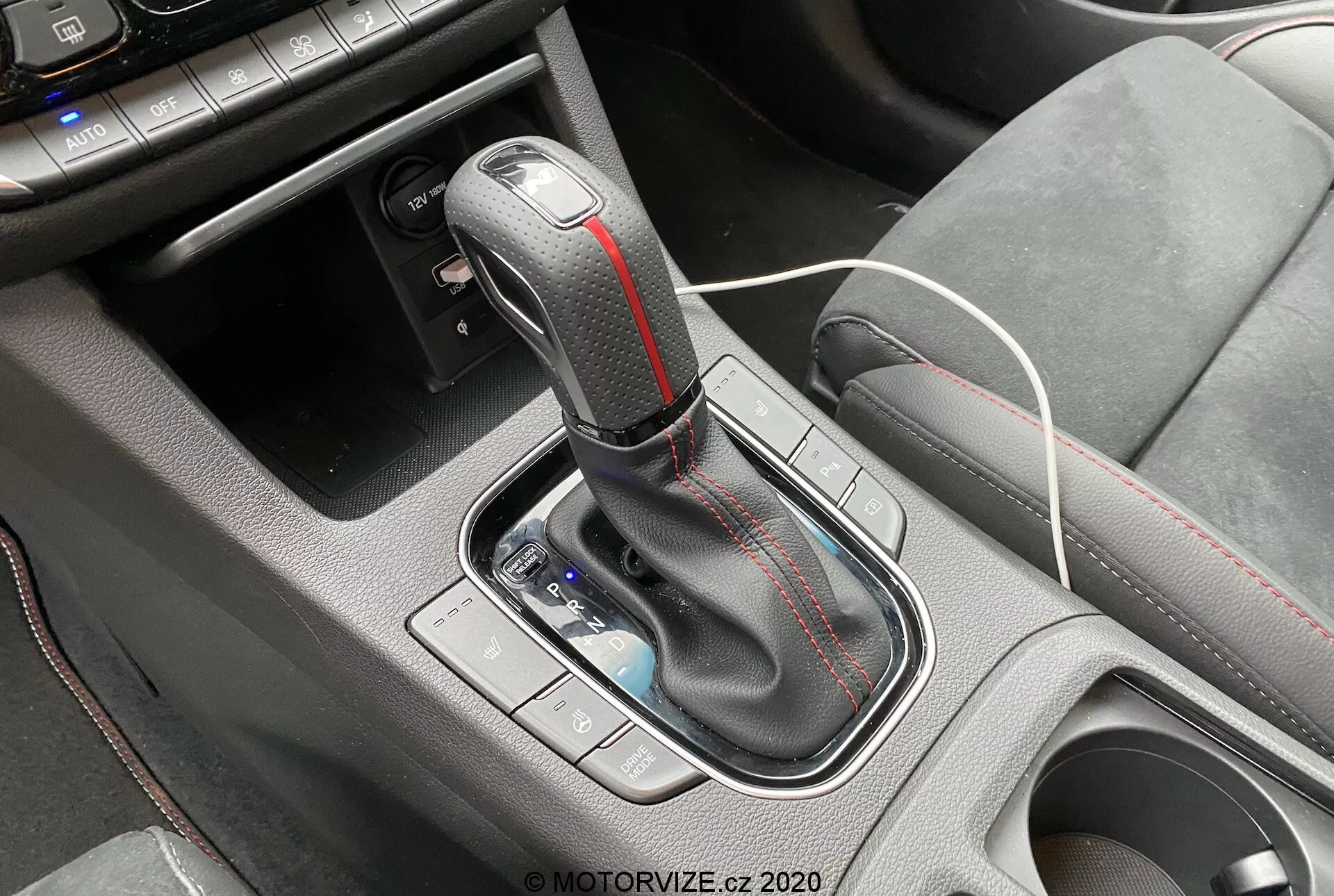 Zbliżenie na obszar konsoli środkowej Hyundai i30 2020 kombi, z automatycznym drążkiem zmiany biegów. Drążek ma gałkę owiniętą skórą z metalicznymi akcentami i czerwonymi kontrastowymi szwami. Przed drążkiem widoczne jest małe schowki z białym kablem do ładowania, co wskazuje na obecność portów USB lub gniazda 12V. Otaczający obszar konsoli wykonany jest z teksturowanego plastiku dla lepszego chwytu. Kontrole po lewej stronie drążka, prawdopodobnie do trybów jazdy lub kontroli trakcji, są lekko rozmyte.