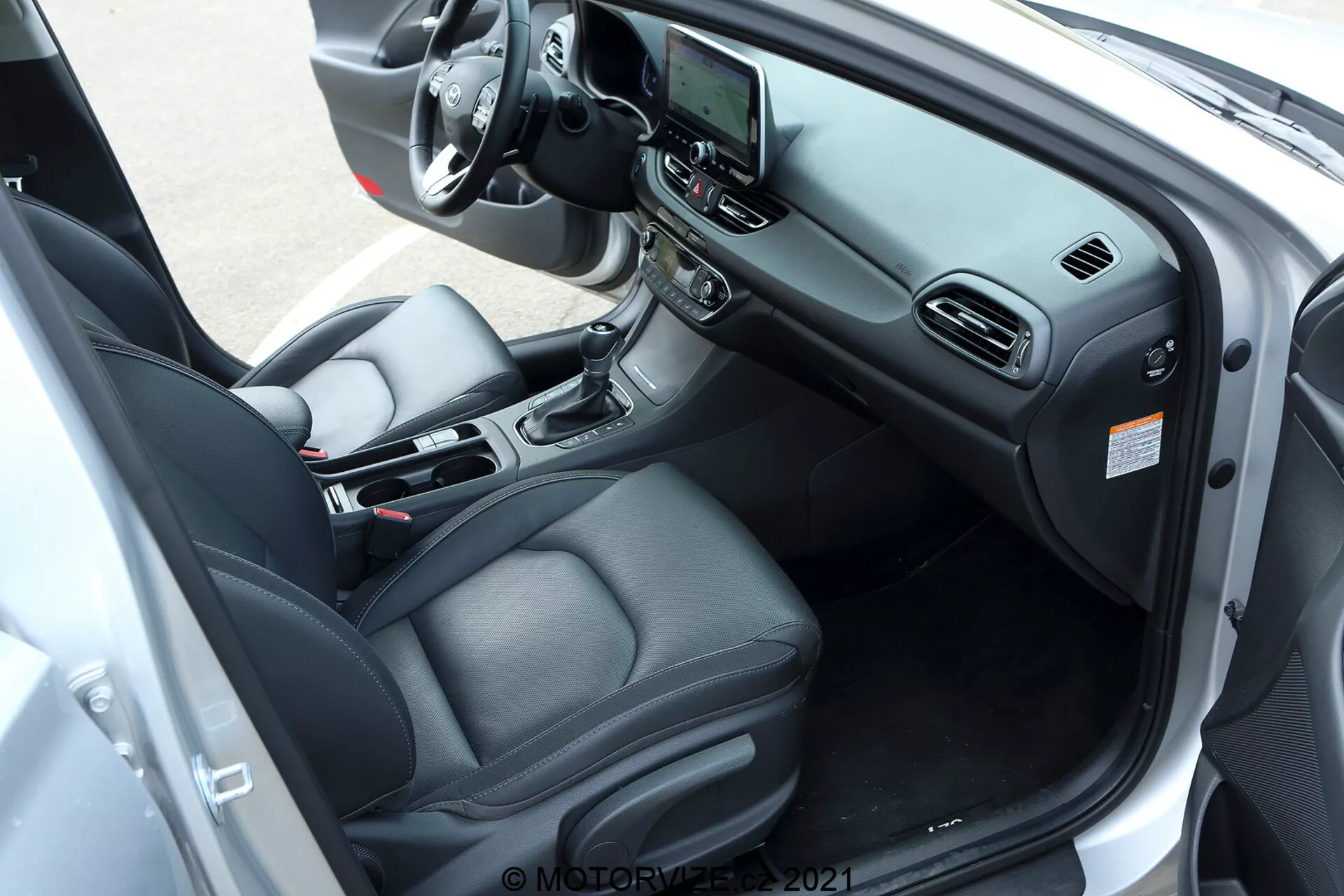 Wnętrze Hyundai i30 kombi 2020 uchwycone z diagonalnego, lekko podwyższonego kąta skupiającego się na stronie kierowcy. Obraz przedstawia pokryty skórą fotel kierowcy z elektronicznymi elementami regulacji, wielofunkcyjną kierownicę z różnymi przyciskami kontrolnymi, centralny system infotainmentowy z ekranem dotykowym oraz automatyczny drążek zmiany biegów na konsoli środkowej. Inne elementy to uchwyty na kubki, schowek, automatyczna klimatyzacja oraz sterowanie elektrycznymi szybami i lusterkami bocznymi na panelu drzwi kierowcy. Drzwi samochodu są szeroko otwarte, aby zapewnić kompleksowy widok na kokpit, układ siedzeń oraz elementy technologii i komfortu wnętrza.