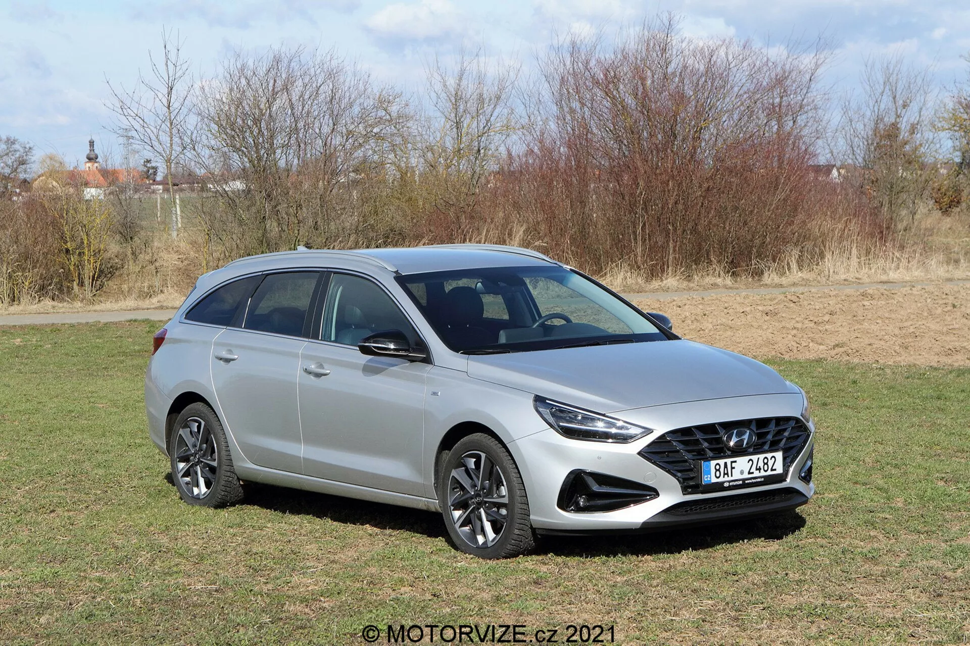 Trójkątny widok z przodu nowoczesnego hatchbacka Hyundai i30 2020 kombi, ujęty pod podwyższonym kątem, ukazujący przedni grill z wzorem plastra miodu, eleganckie reflektory, wieloszprychowe felgi aluminiowe, lusterka boczne z zintegrowanymi kierunkowskazami i relingi dachowe na metalowym nadwoziu z gładkimi, rzeźbionymi liniami w słonecznym otoczeniu naturalnym.