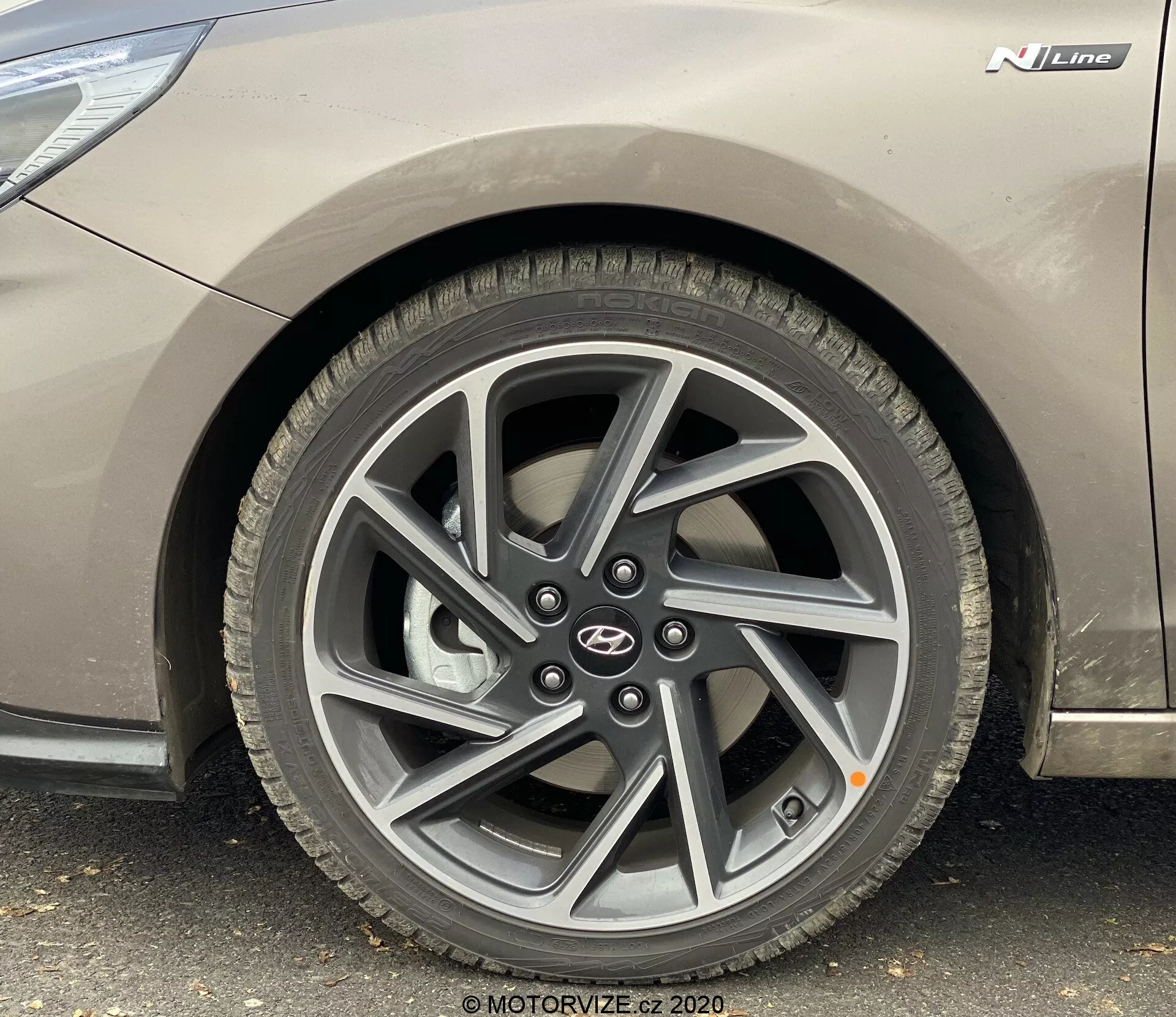 Zbliżony widok z boku z niskiego kąta na przednie koło po stronie kierowcy Hyundai i30 2020 (i30-kombi). Obraz podkreśla wieloramienną felgę w ciemnym kolorze, bieżnik standardowej opony drogowej, metaliczne szare nadwozie pojazdu z odznaką 'N Line' i emblematem producenta.