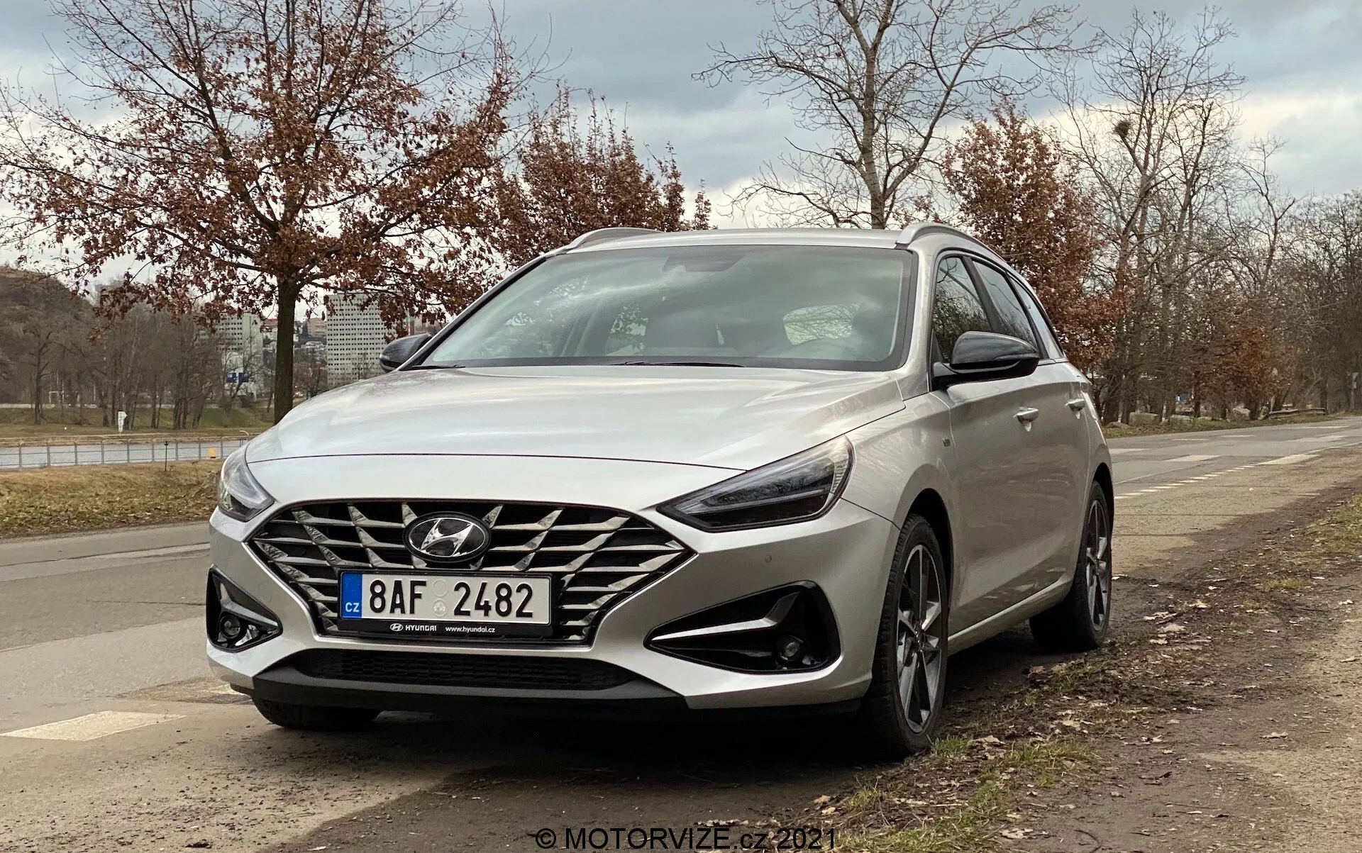 Obraz przedstawia srebrnego Hyundai i30 Kombi 2020 uchwycony pod niskim kątem trzy czwarte od przodu, prezentując projekt z perspektywy kierowcy. Kąt uwypukla dużą kaskadową kratę z wyraźnym logo Hyundai, nowoczesne reflektory LED, wieloramienne felgi, zintegrowane światła do jazdy dziennej poniżej reflektorów oraz widoczny radar do funkcji bezpieczeństwa. Podkreślone są również aerodynamiczne linie i kontury samochodu, z lusterkami bocznymi z wbudowanymi kierunkowskazami. Tło obejmuje krawężnik, drzewa i pochmurne niebo, co sugeruje podmiejskie otoczenie na świeżym powietrzu.