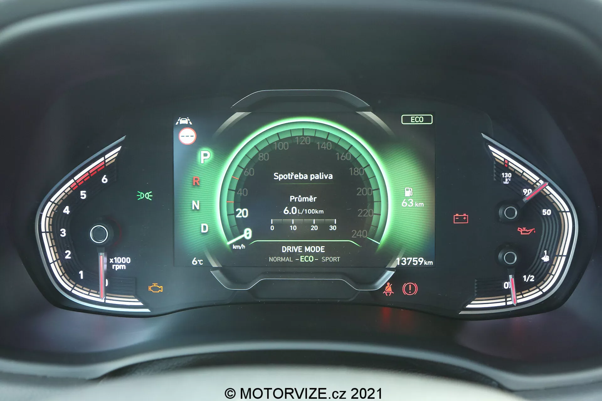 Zbliżenie nowoczesnego zestawu wskaźników Hyundai i30 2020 z perspektywy siedzenia kierowcy, z centralnym wyświetlaczem cyfrowym otoczonym wskaźnikami analogowymi. Po lewej stronie znajduje się obrotomierz, a po prawej wskaźnik poziomu paliwa i temperatury silnika. Różne wskaźniki i ikony pokazują status pojazdu, takie jak zapięcie pasów, naładowanie akumulatora i system hamulcowy. Centralny wyświetlacz zawiera dane dotyczące zużycia paliwa, ustawienia trybu jazdy i odczyt licznika kilometrów. Zestaw wskaźników jest podświetlany na czerwono i zielono, co sugeruje zaawansowany system elektroniczny z możliwymi elementami dotykowymi lub haptycznymi, znak wodny © MOTORVIZE.cz 2021.