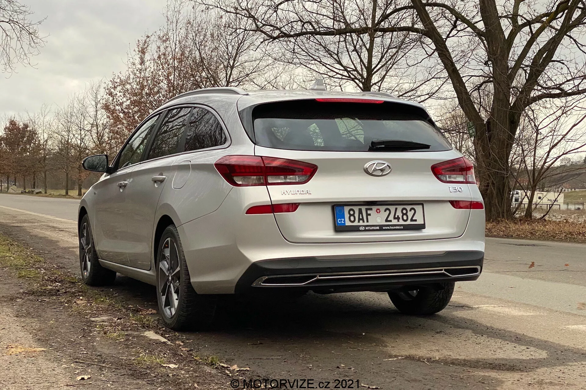 Tylny trójkątny widok srebrnego Hyundaia i30 kombi z 2020 roku, ukazujący elementy tylnego projektu, w tym tylne światła, które być może zawierają technologię LED, tylna wycieraczka, odznaki samochodu i nazwę modelu. Obraz przedstawia również podwójne końcówki wydechu zintegrowane z tylnym zderzakiem, felgi aluminiowe oraz częściowy boczny profil, który sugeruje przestronne wnętrze. Samochód jest zaparkowany na dwupasmowej drodze w obszarze wiejskim lub półwiejski z drzewami i otwartą przestrzenią w tle.