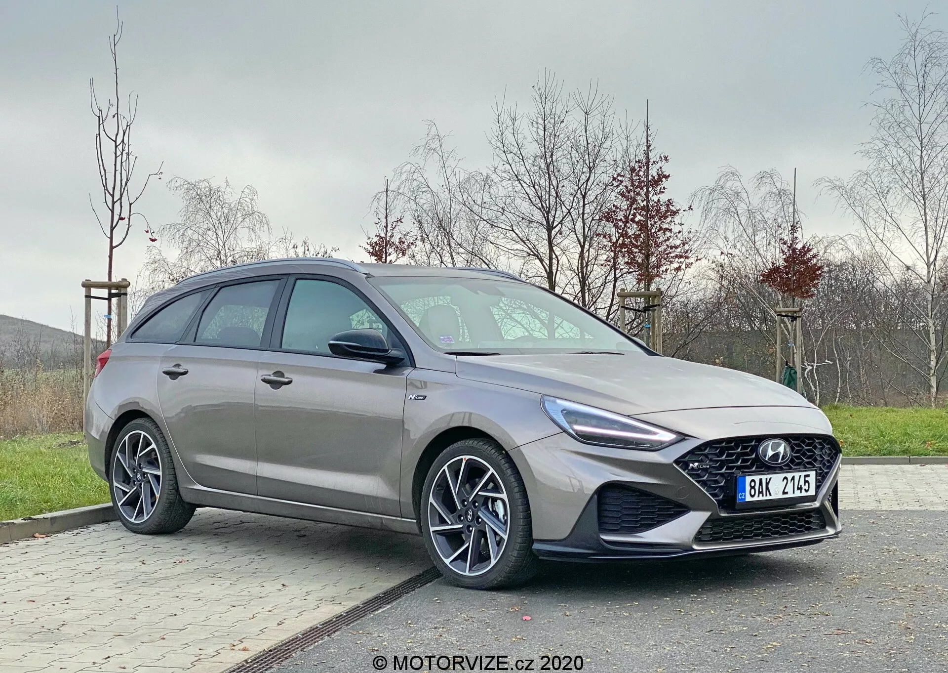 Nowoczesny Hyundai i30 2020 widziany z kąta trzech czwartych przodu. Skupienie na stronie kierowcy ukazującej eleganckie detale projektu takie jak agresywne reflektory z LED-owymi światłami dziennymi, wyraźny grill, wieloramienne felgi aluminiowe i konturowany profil boczny. Ustawiony poza centrum na utwardzonym terenie z trawiastym tłem, przedstawiający dynamiczny, nowoczesny hatchback z zaawansowaną technologią motoryzacyjną.
