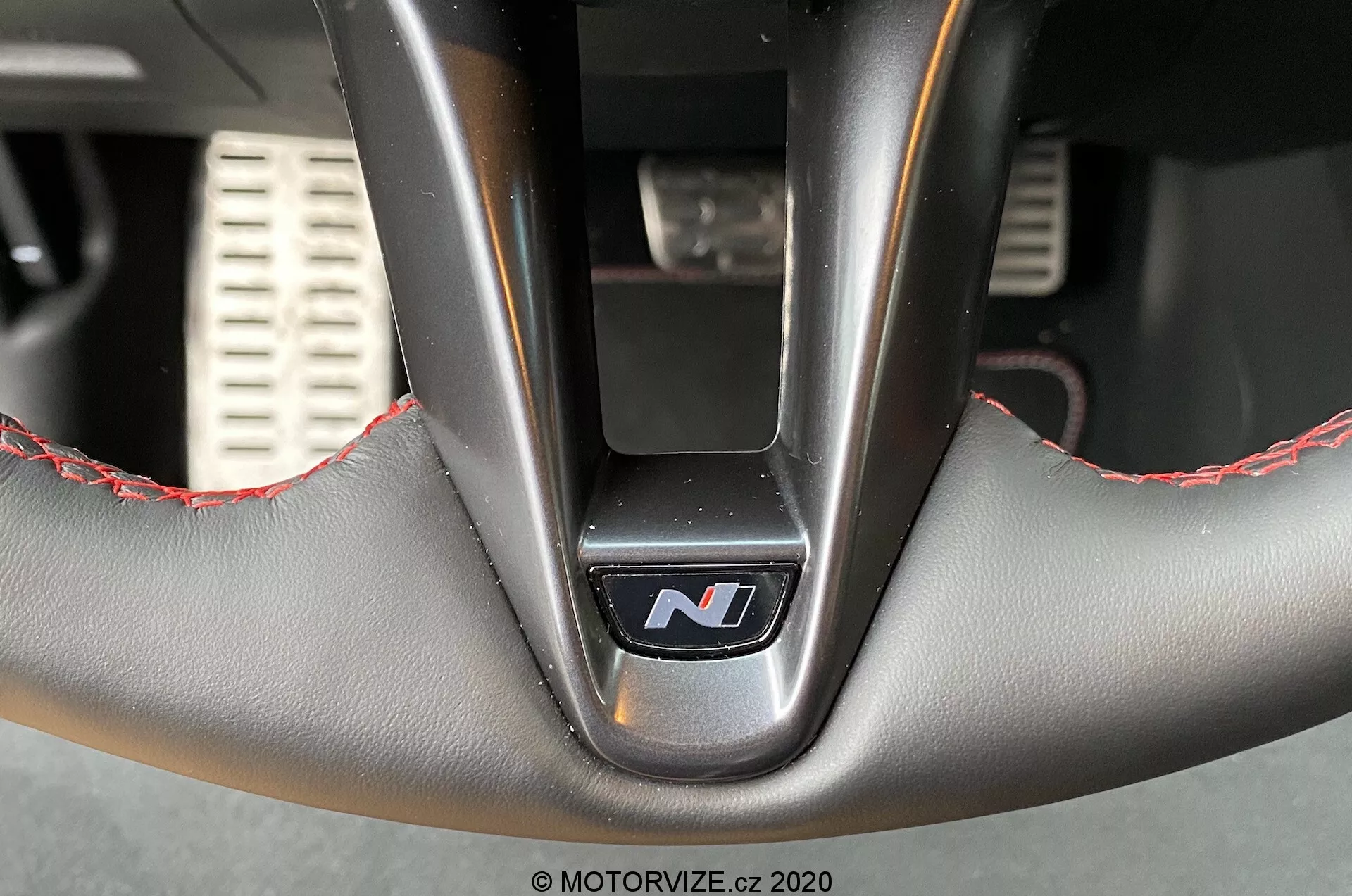Zbliżone wnętrze kierownicy Hyundai i30 2020 (i30-kombi) od dołu, podkreślając dolną część z centralnym emblematem 'N' wskazującym na wersję sportową, skórzaną tapicerką z czerwonymi przeszyciami, metalowymi ramionami i elementami perforowanych sportowych pedałów w nieostrym tle.