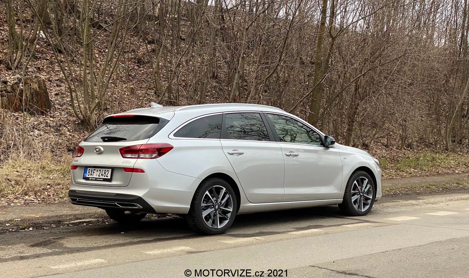 Hyundai i30 2020 w wersji kombi zaparkowany przy miejskiej drodze z widokiem zarówno na profil boczny, jak i tylny, z tylnym spojlerem dachowym, srebrnymi, wieloramiennymi felgami aluminiowymi, podwójnymi rurami wydechowymi zintegrowanymi z tylnym zderzakiem, tylnymi światłami rozciągającymi się na klapę bagażnika, relingami dachowymi i bocznymi lusterkami z wbudowanymi kierunkowskazami. Nowoczesna technologia pojazdu może obejmować czujniki parkowania, kamerę cofania i tylne światła LED. Zdjęcie jest dobrze oświetlone i umożliwia szczegółową obserwację projektu i funkcji samochodu w miejskim środowisku.