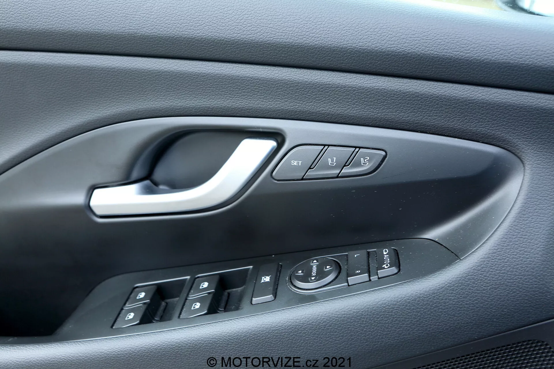 Zbliżenie na panel drzwi kierowcy w Hyundai i30 Kombi 2020, podkreślające skomplikowany design i funkcjonalność, w tym ergonomiczny srebrny uchwyt, cztery przyciski sterowania oknami elektrycznymi z wskazaniem automatycznego opuszczania, elektroniczny joystick do regulacji lusterek bocznych, przyciski blokady i odblokowywania oraz przyciski pamięci fotela. Teksturowany plastikowy panel dodaje estetyki i przyczepności, wskazując na wnętrze bogate w funkcje.