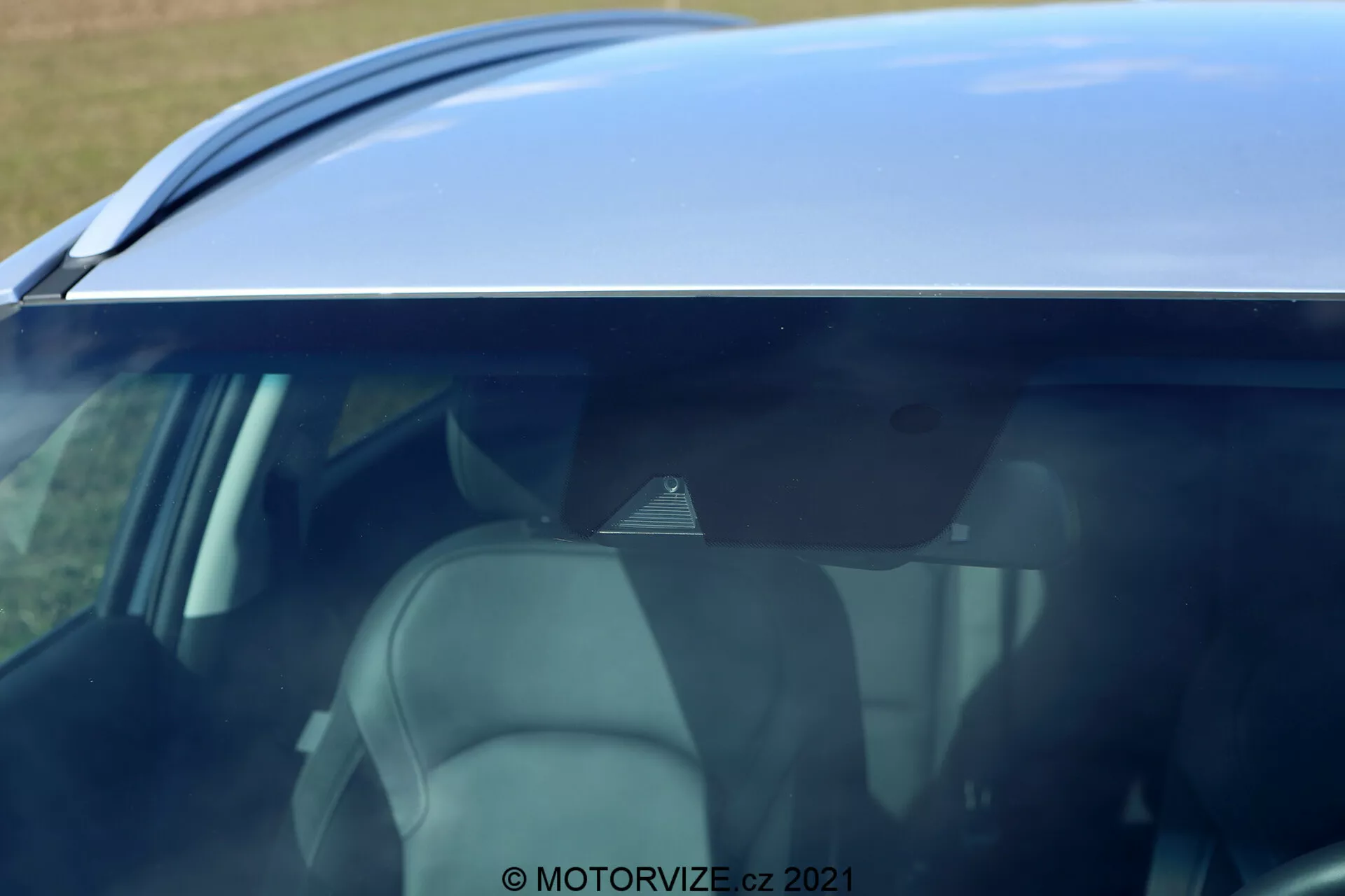 Zbliżenie, kadr pod kątem skupiający się na górnej połowie bocznego profilu Hyundai i30 2020. Widok boczny z podwyższonej perspektywy, ukazujący dach samochodu, górną część drzwi i przyciemniane szyby. Linia dachu posiada gładki, zakrzywiony kształt prowadzący do tylnego spojlera lub anteny typu płetwa rekina. Częściowy widok na boczne szyby ukazuje jasne tapicerki siedzeń, zagłówków i części kabiny pasażerskiej. Odbicie nieba na dachu i szybach wskazuje na jasne otoczenie. Znak wodny © MOTORVIZE.cz 2021 sugeruje, że zdjęcie zostało wykonane w celach przeglądowych lub promocyjnych.