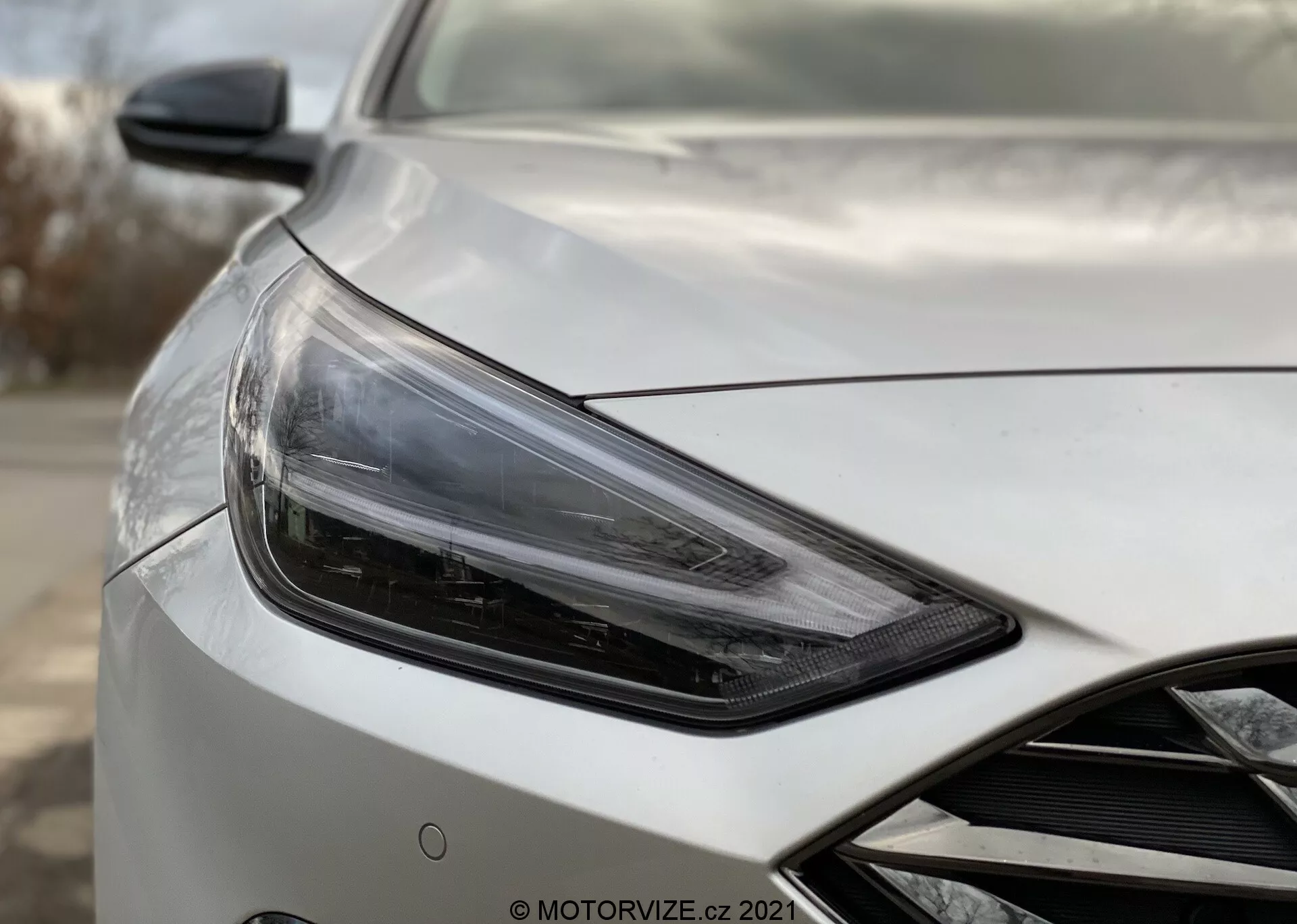 Zbliżenie Hyundai i30 2020 Kombi, przedstawiające przednią lewą sekcję pojazdu pod kątem trzech czwartych z widokiem na stronę kierowcy, z koncentrującym się na eleganckim, kanciastym zestawie reflektorów z nowoczesną technologią LED lub HID, towarzyszącym przez opływowe lusterko boczne z potencjalnymi zaawansowanymi technologiami, takimi jak regulacja elektryczna czy monitorowanie martwych stref. Nadwozie samochodu ma wysokiej jakości perłową lub metaliczną powłokę z aerodynamicznymi konturami na masce i zarysowaniu przedniego grilla. Tło jest rozmyte, aby podkreślić szczegóły samochodu.