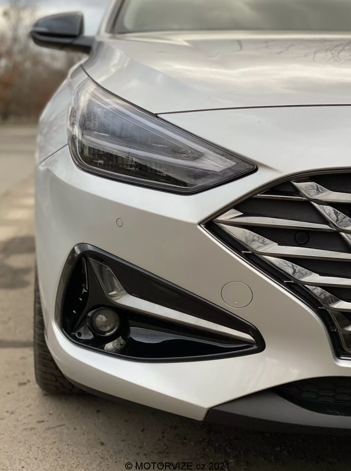 Zbliżenie pod kątem przedniej lewej strony nowoczesnego Hyundai i30 Kombi 2020, koncentrujące się na reflektorze i obszarze zderzaka przedniego. Zdjęcie wykonane z niskiego punktu widzenia w pobliżu przedniego koła, uchwyca kanciasty reflektor LED, chromowaną kratkę, agresywny design zderzaka z dużym wlotem powietrza oraz światłem przeciwmgielnym osadzonym w czarnym wykończeniu. Metaliczne wykończenie lakieru odbija światło, a linie wzornicze na masce podkreślają dynamiczny wygląd samochodu. Tło jest rozmyte, aby uwydatnić szczegóły pojazdu, sugerując zaawansowane funkcje, takie jak adaptacyjne oświetlenie i potencjalne czujniki parkowania.