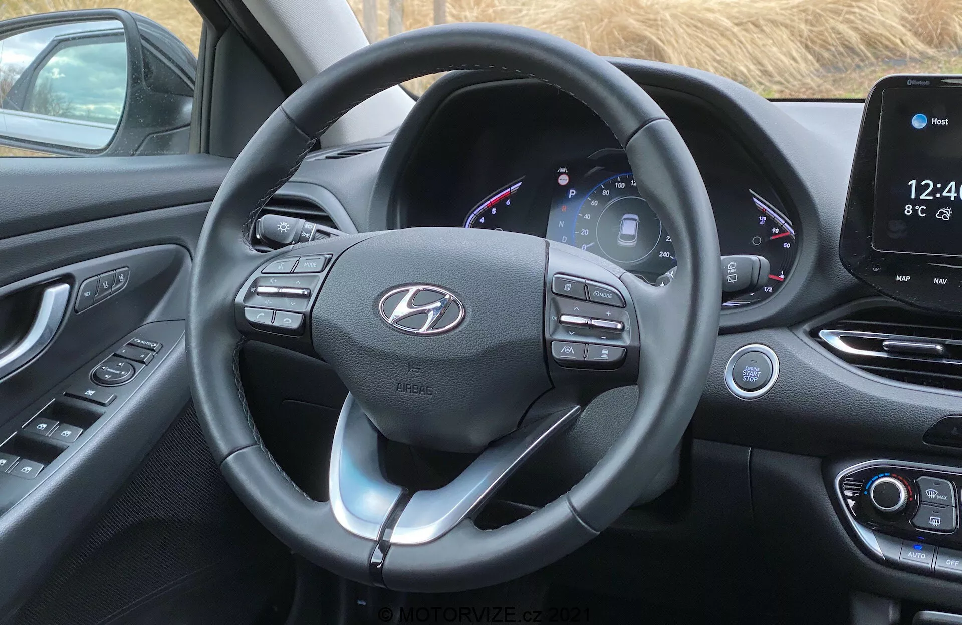 Obraz wnętrza przedniej kabiny Hyundai i30 2020 z perspektywy kierowcy. Skupia się na nowoczesnej kierownicy z wieloma przyciskami sterującymi i łopatkami zmiany biegów, skórzanym obiciem ze widocznymi szwami oraz centralnym emblematem logo. Za kierownicą znajduje się częściowo cyfrowy zestaw wskaźników wyświetlający prędkościomierz, obrotomierz, wskaźnik paliwa i inne informacje o pojazdu z kolorowym interfejsem. Na prawo, konsola centralna jest wyposażona w system informacyjno-rozrywkowy z ekranem dotykowym wyświetlającym godzinę, temperaturę i interfejs główny, z fizycznymi przyciskami i pokrętłami klimatyzacji poniżej. Kabina charakteryzuje się nowoczesnym designem z zakrzywionymi powierzchniami, miękkimi w dotyku materiałami i czarno-srebrnym lub chromowanym schematem kolorystycznym, podkreślającym nowoczesny i wyszukany wygląd.