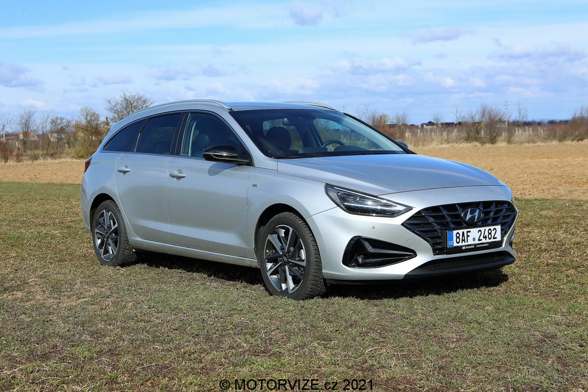 Hyundai i30 Kombi 2020 sfotografowany z lewego przedniego trzykwartego kąta na otwartym polu. Elegancki srebrny hatchback z nowoczesnym aerodynamicznym designem, wyrazistą przednią kratką, ostrymi reflektorami LED, wieloramiennymi felgami aluminiowymi, światłami przeciwmgielnymi, spojlerem dachowym i relingami dachowymi. Tło zewnętrzne z jałowymi drzewami, podkreślające zaawansowaną technologię oświetleniową i możliwe funkcje bezpieczeństwa, takie jak zintegrowane czujniki i kierunkowskazy w lusterkach bocznych.