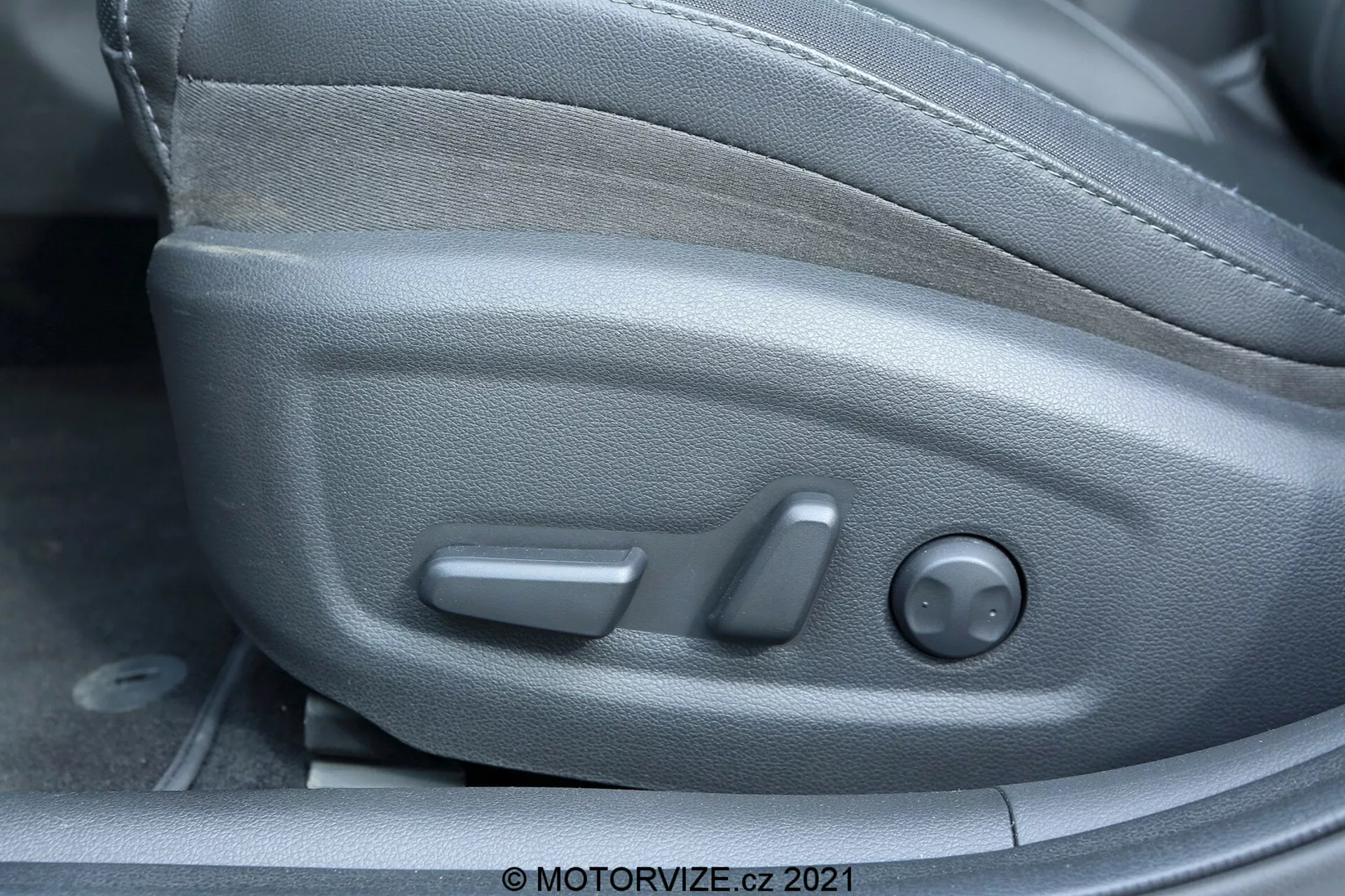 Zbliżenie wnętrza Hyundai i30 2020, koncentrujące się na przyciskach fotela kierowcy z bocznego kąta. Widoczne funkcje obejmują regulacje fotela: przedni chwyt służący do przesuwania, środkową dźwignię do odchylania oraz tylne, okrągłe pokrętło do regulacji wysokości. Plastikowa obudowa integruje się z bocznym stylem fotela, a tapicerka z tkaniny zapewnia trwałość i komfort.