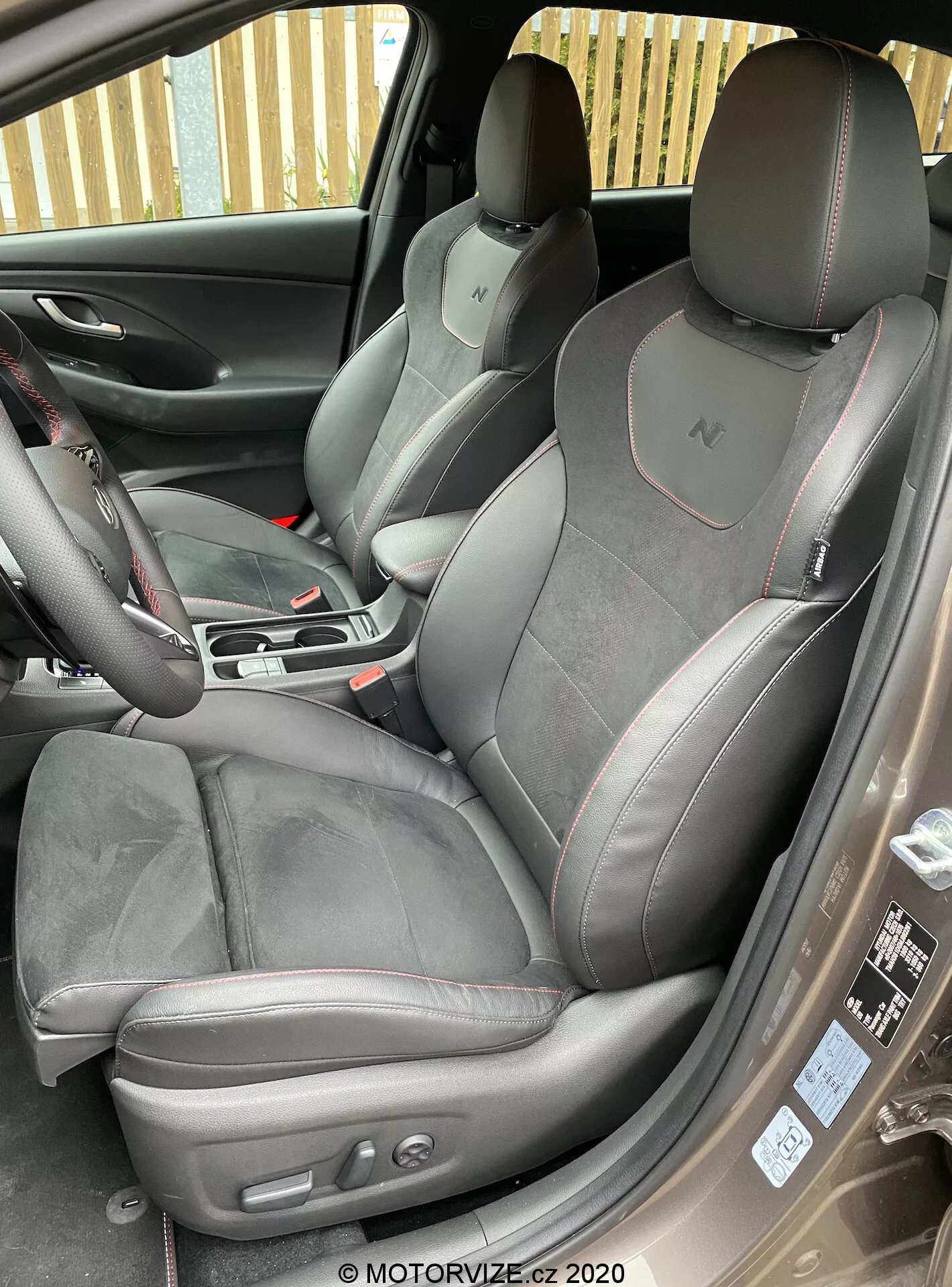 Ten szczegółowy obraz przedstawia wnętrze przedniej kabiny Hyundai i30 2020 (i30-kombi) z lekko uniesionego kąta, patrząc z zewnątrz otwartego drzwi przedniego pasażera. Otwarta klapa pasażerska ukazuje sportowe siedzenie premium tapicerowane zamszem lub alcantarą i skórą z kontrastowym przeszyciem. Zintegrowane zagłówki z logo 'N' wskazują na poziom wyposażenia samochodu. Siedzenie kierowcy posiada przyciski sterujące foteli elektrycznie sterowanych, a ich układ graficzny uwydatnia nowoczesne technologiczne i komfortowe funkcje samochodu.