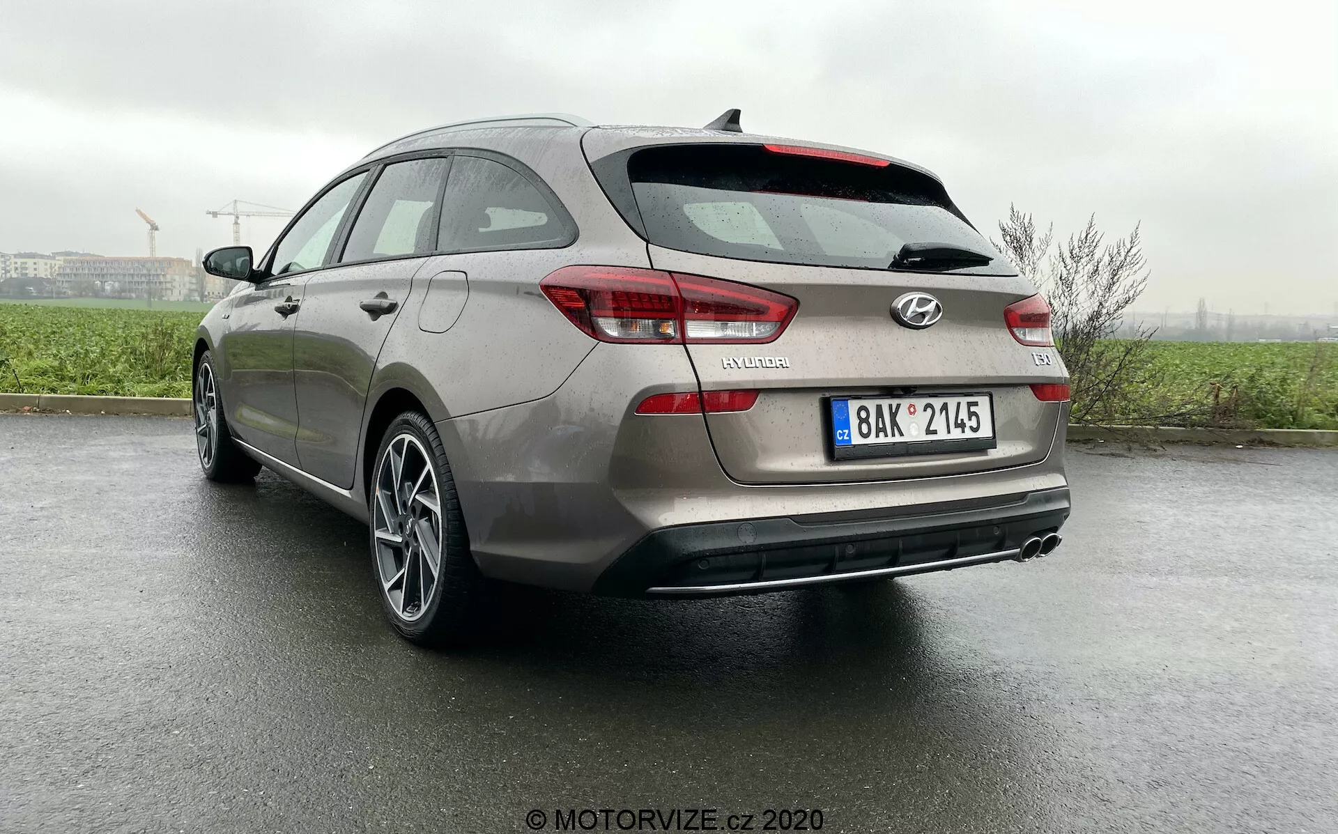 Trzypunktowy widok tylnej części Hyundai i30 Kombi 2020 z niskiego kąta, pokazujący szczegóły designu, w tym tylne światła, tylny zderzak, drzwi bagażnika, kamerę parkowania tylną, felgi aluminiowe i odblaski na mokrej powierzchni po niedawnym deszczu, podkreślając zaawansowane systemy wspomagania kierowcy i estetyczny wygląd pojazdu.