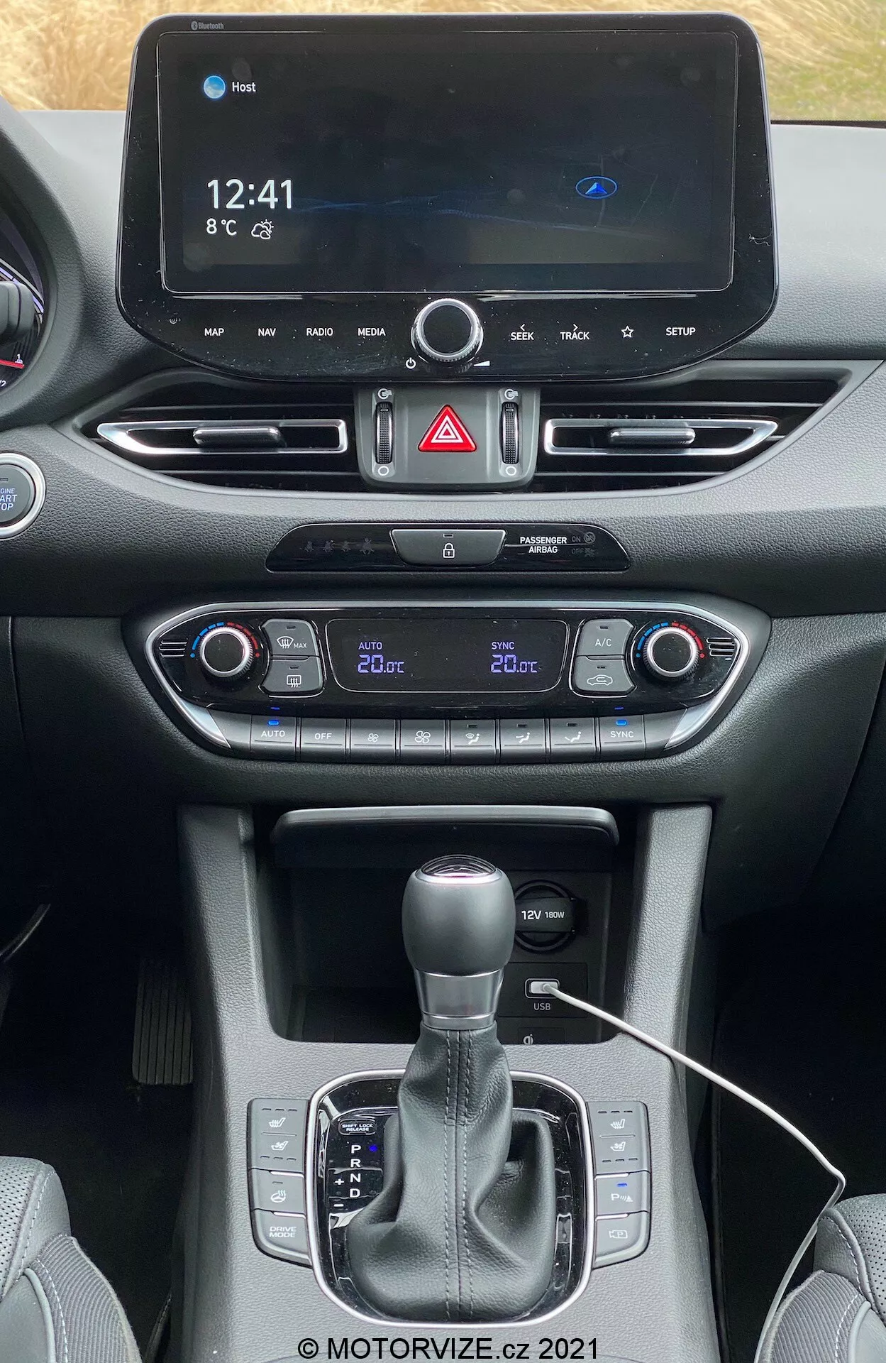 Prosty widok na centralną konsolę Hyundai i30 Kombi 2020 z foteli przednich, pokazujący cyfrowy system infotainment wyświetlający czas (12:41), temperaturę (8°C) i ikonę Bluetooth. Poniżej znajduje się przycisk ostrzegawczy, dwustrefowa klimatyzacja z osobnymi ustawieniami temperatury (kierowca 22°C, pasażer 20°C), przycisk start-stop, dźwignia zmiany biegów automatycznych z trybem manualnym, potencjalne uchwyty na kubki/przestrzeń na przechowywanie, port łączności z widocznym kablem i mieszane materiały kokpitu oraz konsoli. Na dole znacznik wodny z prawami autorskimi '@ MOTORVIZE.cz 2021'.