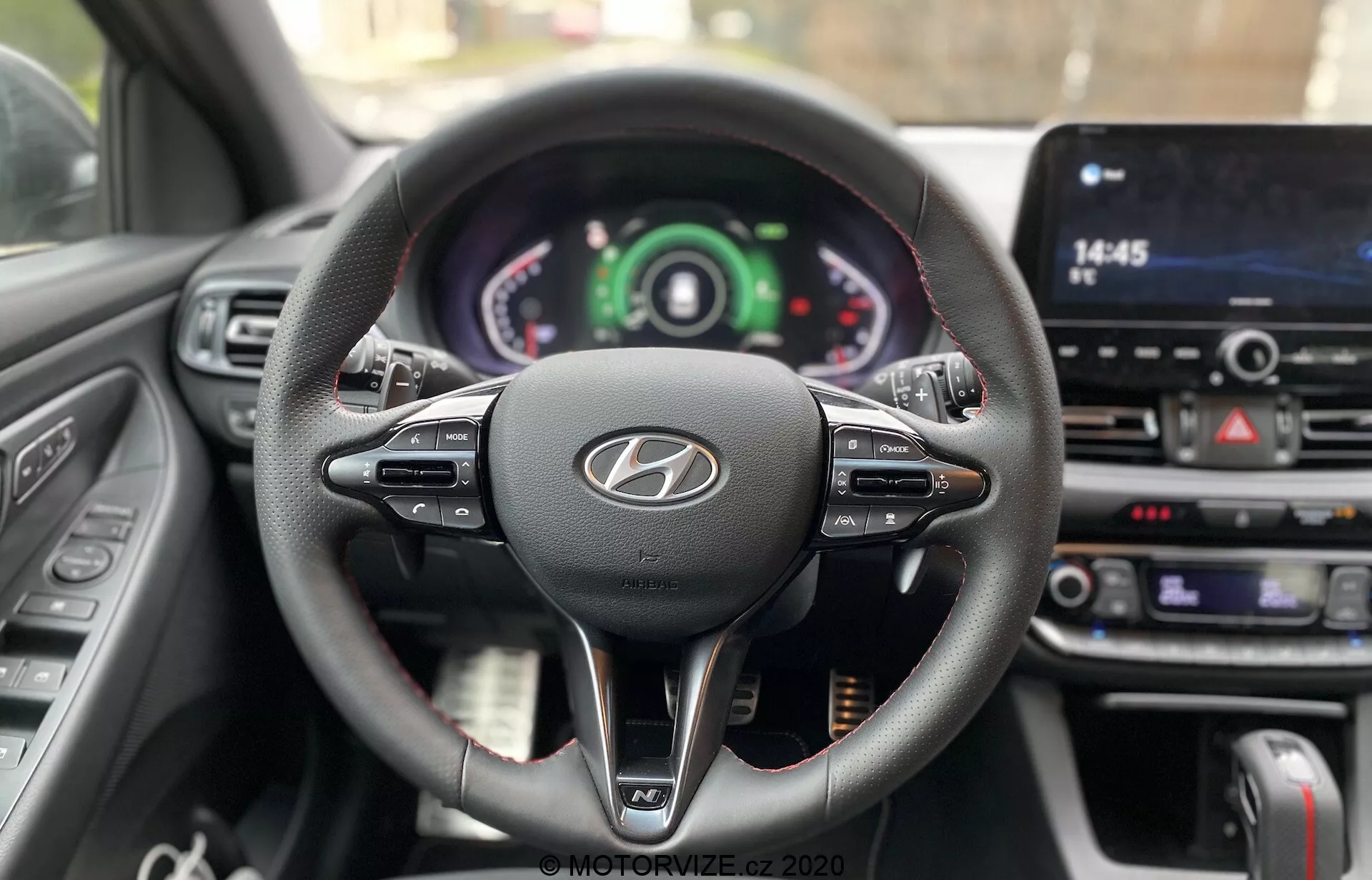 Widok z perspektywy kierowcy na kierownicę i deskę rozdzielczą Hyundai i30 2020, pokazujący trójramienną, skórzaną konstrukcję z multifunkcyjnymi przyciskami, wyraźnym logo Hyundai oraz zestawem wskaźników z analogowymi i cyfrowymi wyświetlaczami. Centralnie umieszczony ekran dotykowy infotainmentu i przyciski klimatyzacji podkreślają zaawansowane technologie samochodu i elementy interaktywne kierowcy.