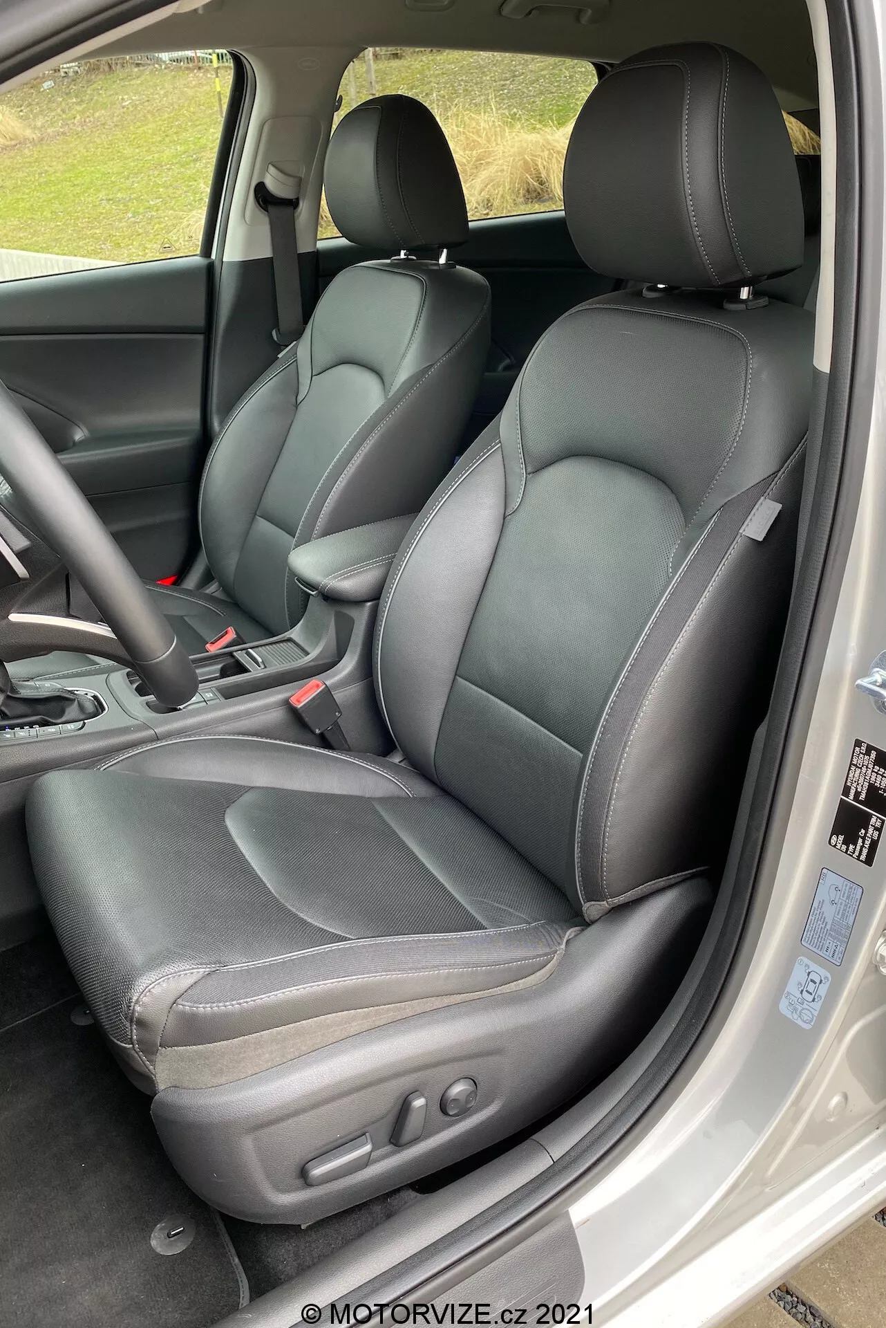 Szczegółowy widok wnętrza przedniej części Hyundai i30 Kombi 2020, skupiający się na stronie kierowcy. Perspektywa jest nieco powyżej poziomu siedzenia, patrząc ukośnie w dół do kabiny, jakby wchodząc przez tylne drzwi. Czarny skórzany fotel kierowcy jest wyraźnie widoczny z widocznymi szwami i elektronicznymi kontrolkami regulacji z boku. Zauważalne są również pasy bezpieczeństwa i lewa trzecia część trójramiennej kierownicy. Części konsoli centralnej, w tym pionowa dźwignia zmiany biegów automatyczna w pozycji ‘park’, oraz części deski rozdzielczej i nawiewy powietrza są częściowo widoczne. Kabina prezentuje jednolitą czarną kolorystykę, z różnorodnością faktur na siedzeniach, panelach drzwiowych i desce rozdzielczej. Zdjęcie jest dobrze oświetlone światłem naturalnym, co sugeruje zachmurzone lub zacienione otoczenie.
