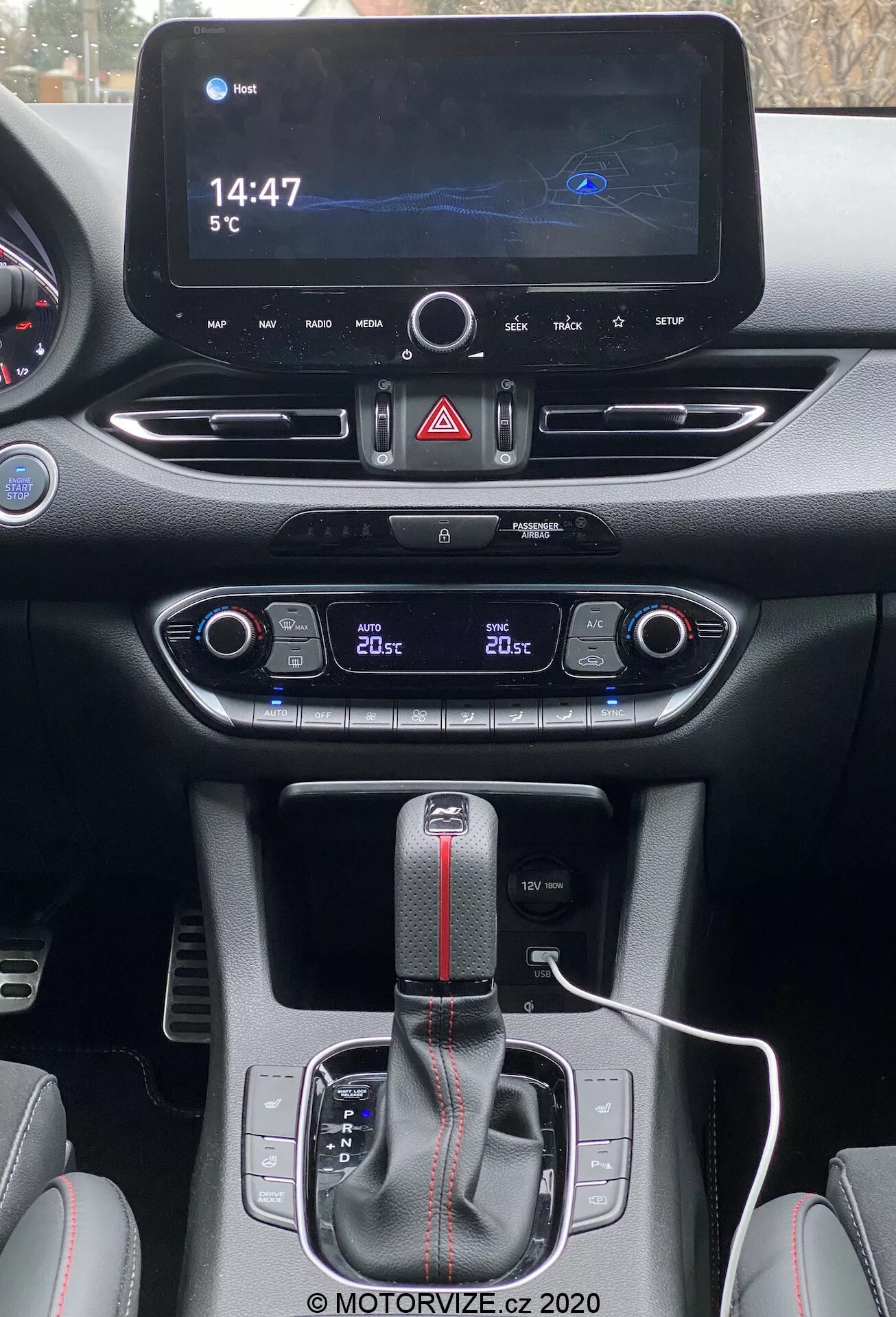 Widok z perspektywy kierowcy na centralną konsolę Hyundai i30 Kombi 2020. Na górze nowoczesny ekran infotainment wyświetla mapę nawigacji, zegar cyfrowy wskazujący '14:47' i temperaturę zewnętrzną '5°C'. Poniżej znajdują się przycisk świateł awaryjnych i kratki wentylacyjne. Niżej panel sterowania klimatyzacją dwustrefową pokazuje '20°C' po obu stronach. Dźwignia automatycznej skrzyni biegów z czarnym mieszkiem i czerwonym paskiem sugeruje sportowe wykończenie. Biały kabel do ładowania jest podłączony koło dźwigni zmiany biegów, otoczony przyciskami prawdopodobnie do wyboru trybów jazdy lub kontroli trakcji. Materiały prezentują gładkie i teksturowane powierzchnie z kontrastowymi przeszyciami, podkreślając nowoczesny, sportowy wystrój wnętrza.