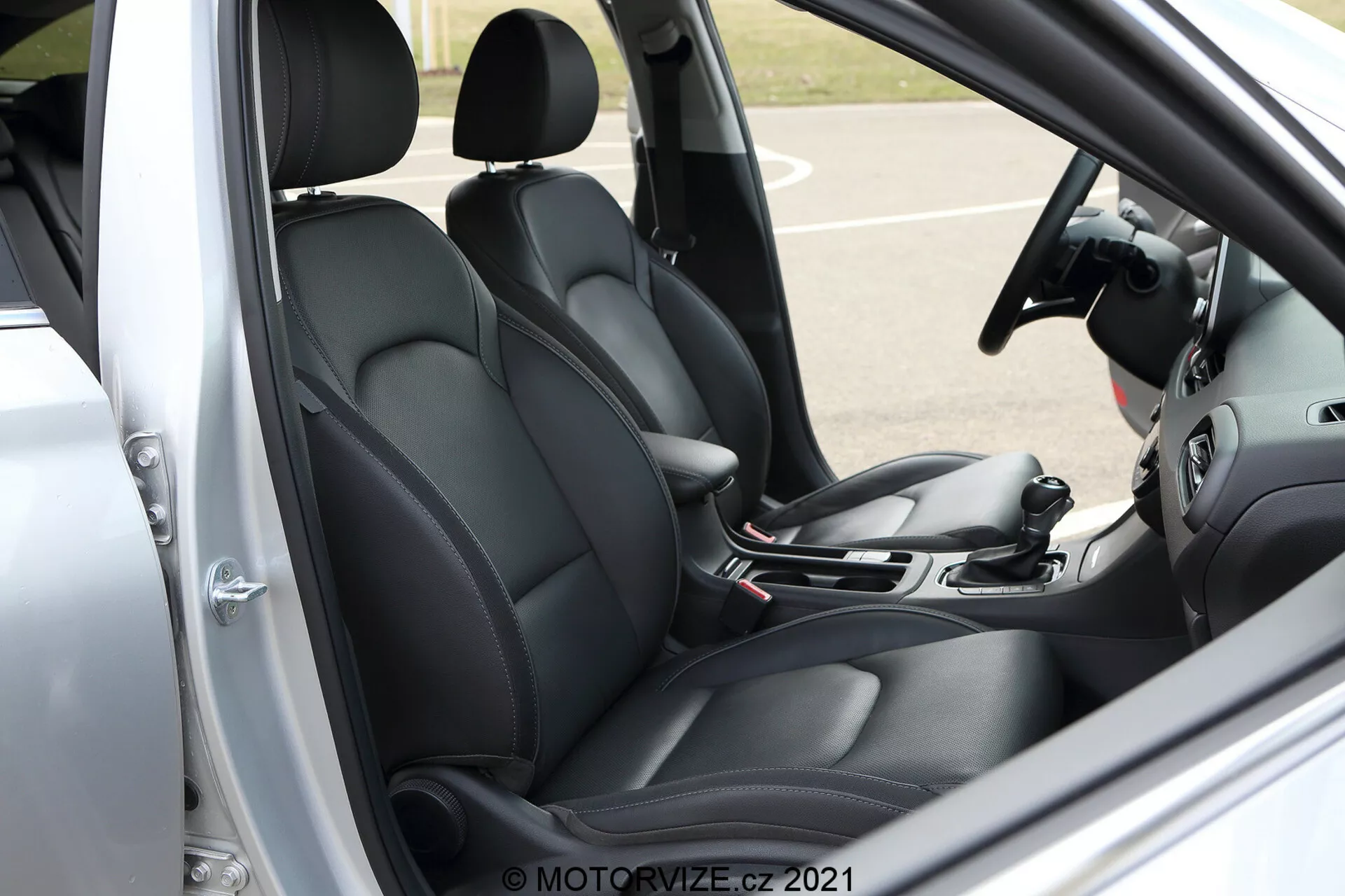 Zbliżenie pod kątem na wnętrze przedniej kabiny Hyundai i30 Kombi 2020. Obraz przedstawia fotel kierowcy i część fotela pasażera, oba tapicerowane na czarno z profilowanym designem dla większego komfortu i wsparcia. Widoczny z boku kierowcy z otwartymi drzwiami, scena obejmuje dźwignię zmiany biegów, ręczny hamulec i części systemów infotainment i klimatyzacji, sugerując nowoczesne funkcje technologiczne. Panel drzwi kierowcy, pokazujący przyciski sterowania szybami i prawdopodobnie regulację lusterek, dodaje do ogólnego funkcjonalnego wyglądu. Kierownica, chociaż częściowo obcięta, sugeruje zintegrowane kontrolki dla lepszej interakcji z pojazdem. Znak wodny 'MOTORVIZE.CZ 2021' w prawym dolnym rogu.