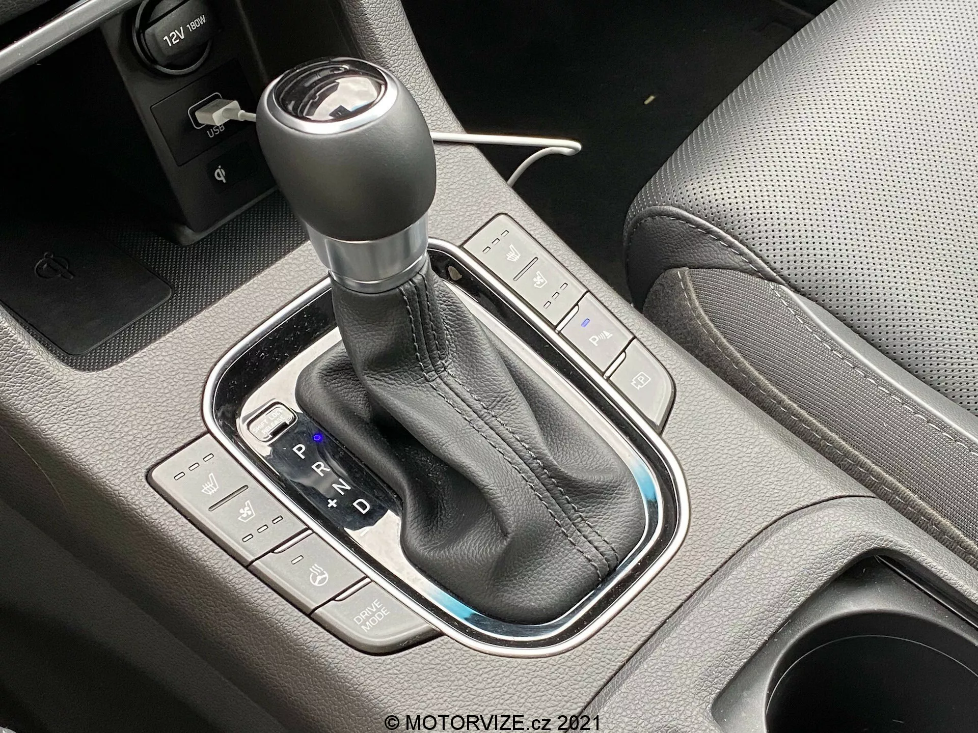 Szczegółowe zbliżenie na automatyczną skrzynię biegów Hyundai i30 2020, zdjęcie wykonane pod kątem 45 stopni. Dźwignia zmiany biegów otoczona jest błyszczącym czarnym obramowaniem, z widoczną skórzaną osłoną i szwami. Układ PRND jest wyraźnie widoczny wraz z oznaczeniami trybu manualnego lub sportowego. Przyciski po lewej stronie kontrolują tryby jazdy, a te po prawej kontrolują stabilność pojazdu lub kontrolę trakcji. Widoczny jest również elektroniczny hamulec postojowy. Dobrze oświetlone zdjęcie podkreśla wysoką jakość i precyzyjne wykonanie centralnej konsoli.