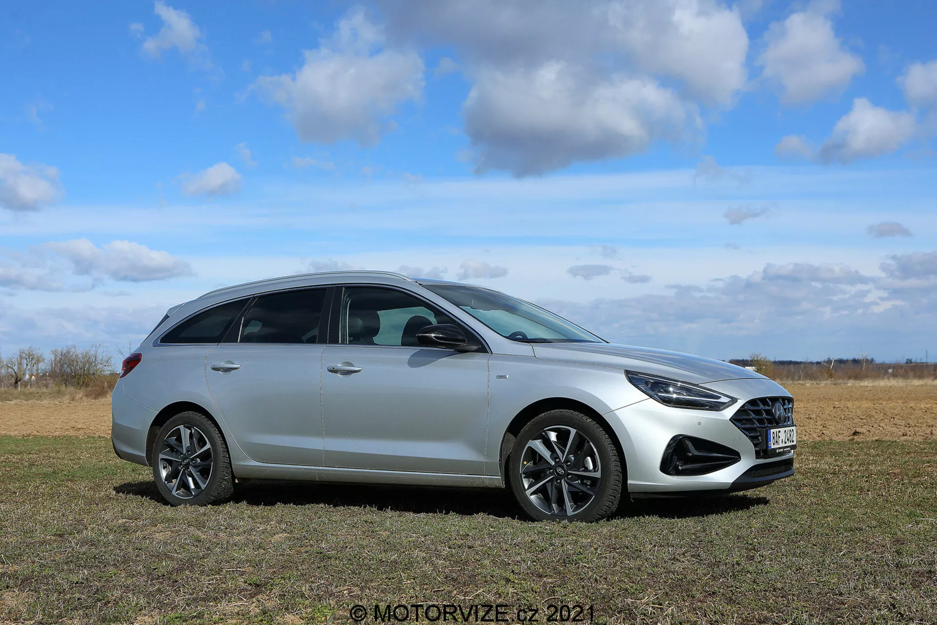 Hyundai i30 2020 Kombi - Nowoczesny samochód typu hatchback na trawiastym polu z czystym niebem w tle, zdjęcie z boku pod kątem przedstawiającym frontowy grill, design kół, reflektory, lusterka boczne, okna, klamki drzwi i linię dachu; wyróżnia się eleganckim metalicznym lakierem, alufelgami oraz zaawansowanymi funkcjami oświetlenia i aerodynamiki.