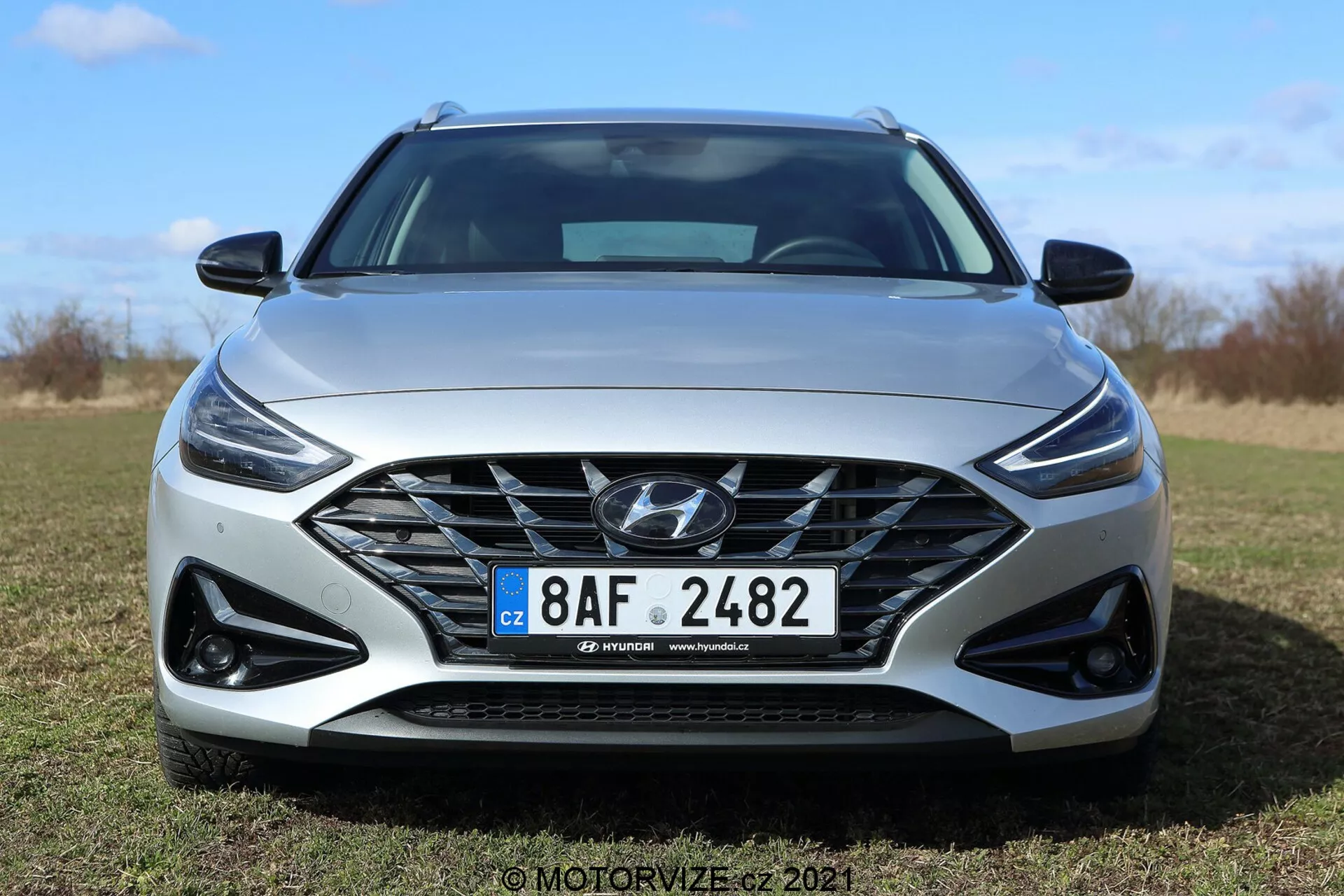 Widok z przodu Hyundaia i30 Kombi 2020 w słoneczny dzień w plenerze. Nowoczesny design samochodu obejmuje dużą kratownicę złożoną z drobnej siatki i logo Hyundai, eleganckie reflektory LED ze światłami dziennymi oraz sportowe obudowy świateł przeciwmgielnych w czerni. Dynamiczny przedni zderzak i wyprofilowana maska podkreślają współczesną i aerodynamiczną estetykę. Widoczna jest europejska tablica rejestracyjna i prawdopodobnie felgi aluminiowe. Jasnoszary kolor samochodu podkreśla jego wyrafinowany styl.