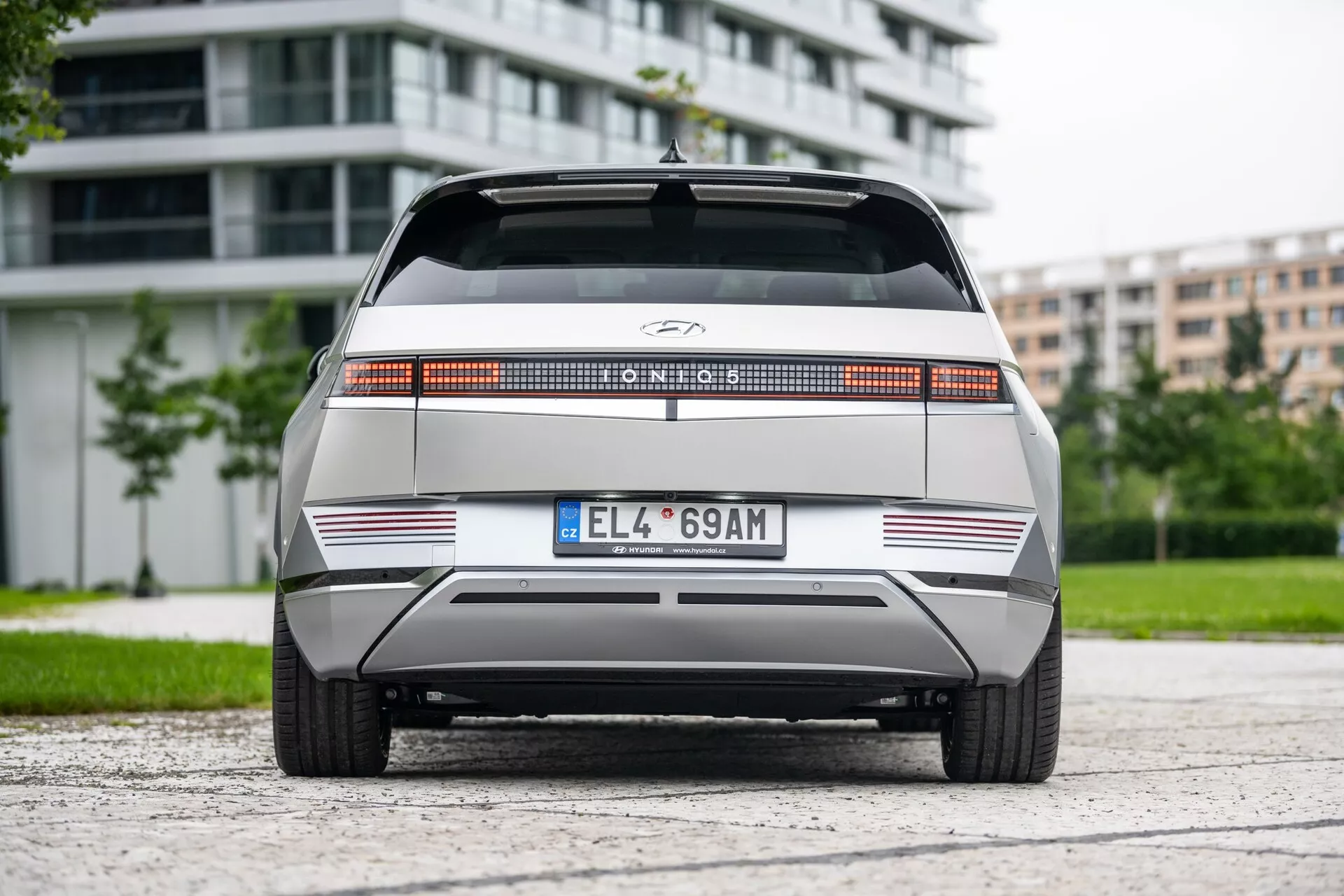 Widok tylni Hyundai Ioniq 5 2WD 2021, z charakterystycznym pełnowymiarowym pasem tylnym, centralnie umieszczonym logo producenta, płaskim tyłem z ostrymi poziomymi liniami, minimalistycznym tylnym zderzakiem z odblaskami oraz europejską tablicą rejestracyjną. Pojazd znajduje się na środku asfaltowej powierzchni, w tle widać zieleń i nowoczesne budynki, oświetlone pochmurnym światłem.