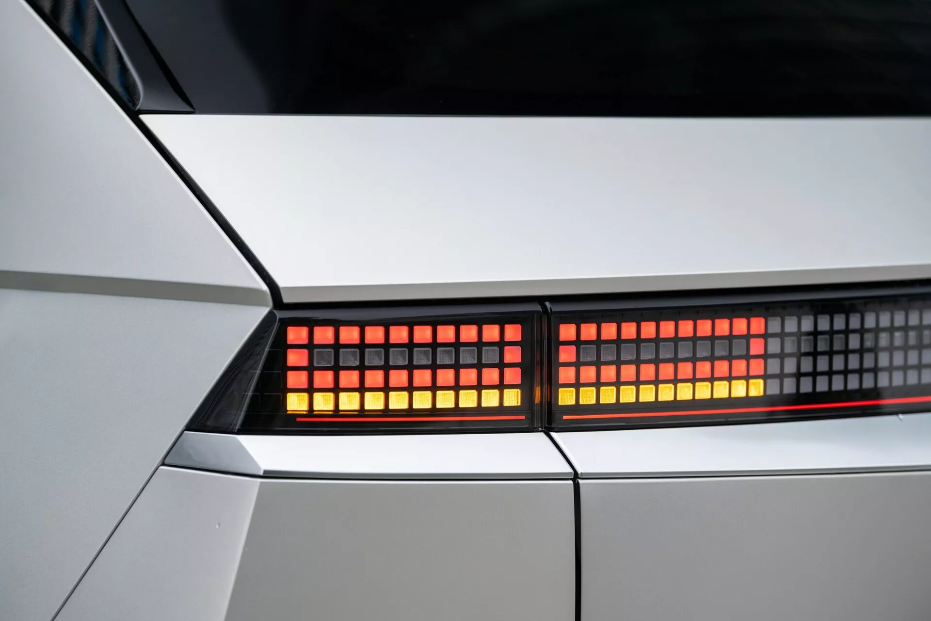 Zbliżenie zmontowanego tylnego światła modelu Hyundai Ioniq-5 2021 2WD. Skupienie na czerwonych, pikselowanych diodach LED w wąskim kadrze, podkreślającym ostrą, geometryczną karoserię w kolorze białym lub jasnosrebrnym. Szczegółowy widok nowoczesnych elementów wzornictwa i technologii oświetleniowej.