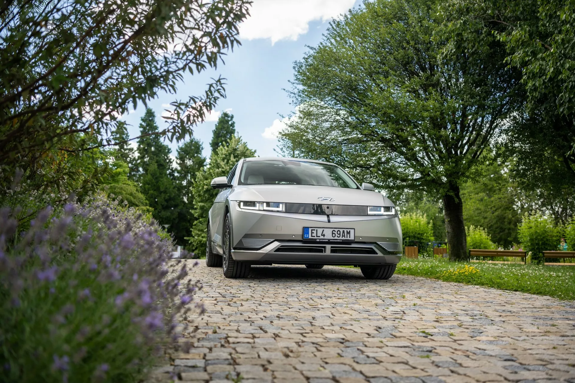 Ujęcie pod niskim kątem Hyundaia Ioniq 5 2WD z 2021 roku zaparkowanego na brukowanej powierzchni. Aparat skupia się na przodzie samochodu, w tym na grillu, lewych reflektorach oraz części maski i przedniej szyby, z tłem zielonych drzew, krzewów i fioletowych kwiatów w dużym świetle dziennym.