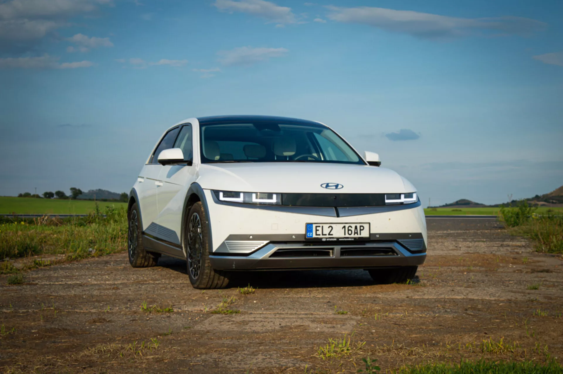 Elektryczny samochód Hyundai Ioniq 5 z 2021 roku prezentujący nowoczesny design z elegancką maską, charakterystycznymi diodowymi światłami do jazdy dziennej, zamkniętym grillem dla aerodynamiki i sportowymi kołami, wykonane z niskiego kąta frontowego, aby podkreślić stylizację i postawę przedniej części samochodu, zaparkowane na zewnątrz pod niebieskim niebem