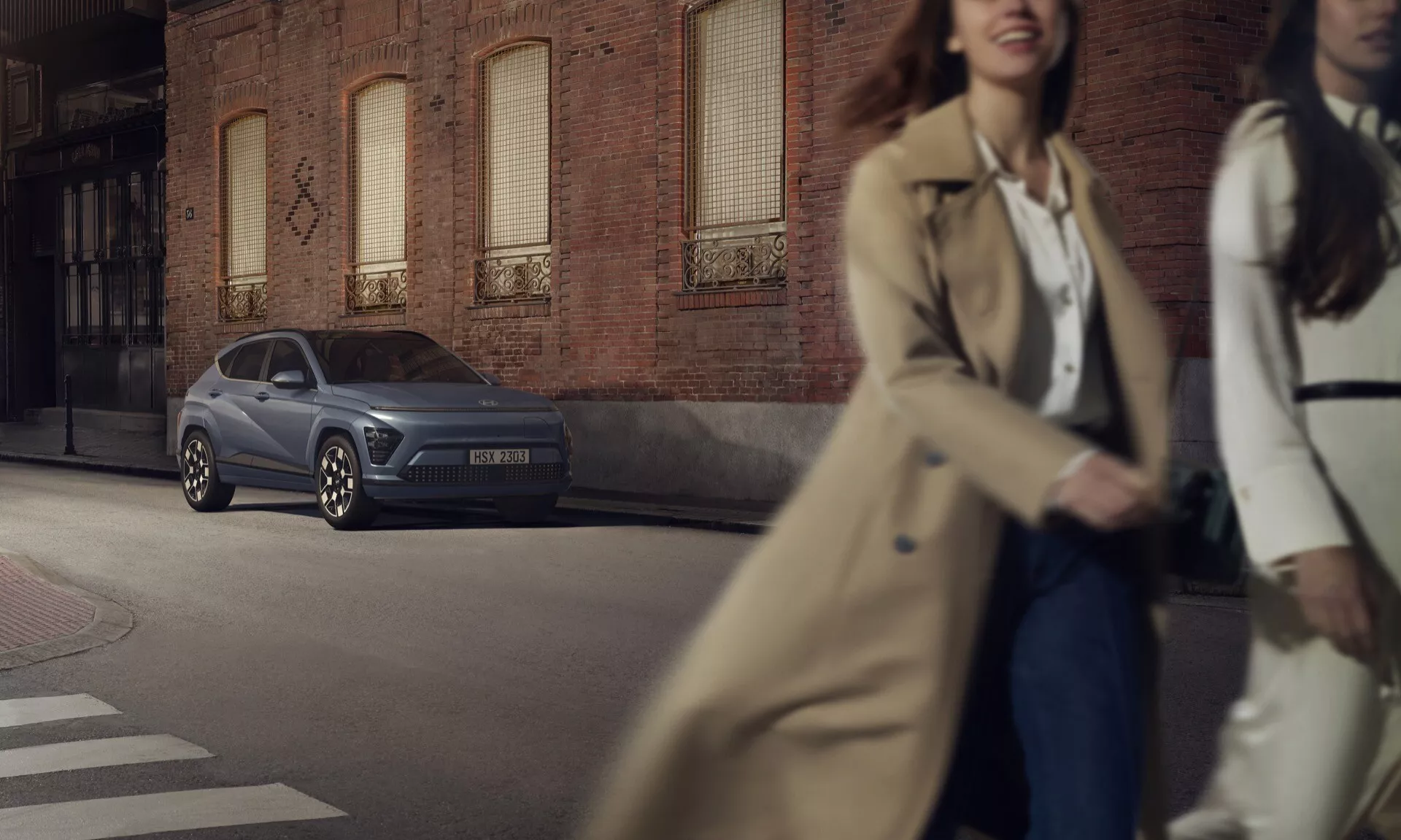 Hyundai Kona 2023 zaparkowany na miejskiej ulicy pod kątem trzech czwartych z przodu, ukazujący eleganckie reflektory, wyraźną atrapę chłodnicy i felgi ze stopu lekkiego o wielu ramionach na tle fasady z cegły z przechodniami w ruchu