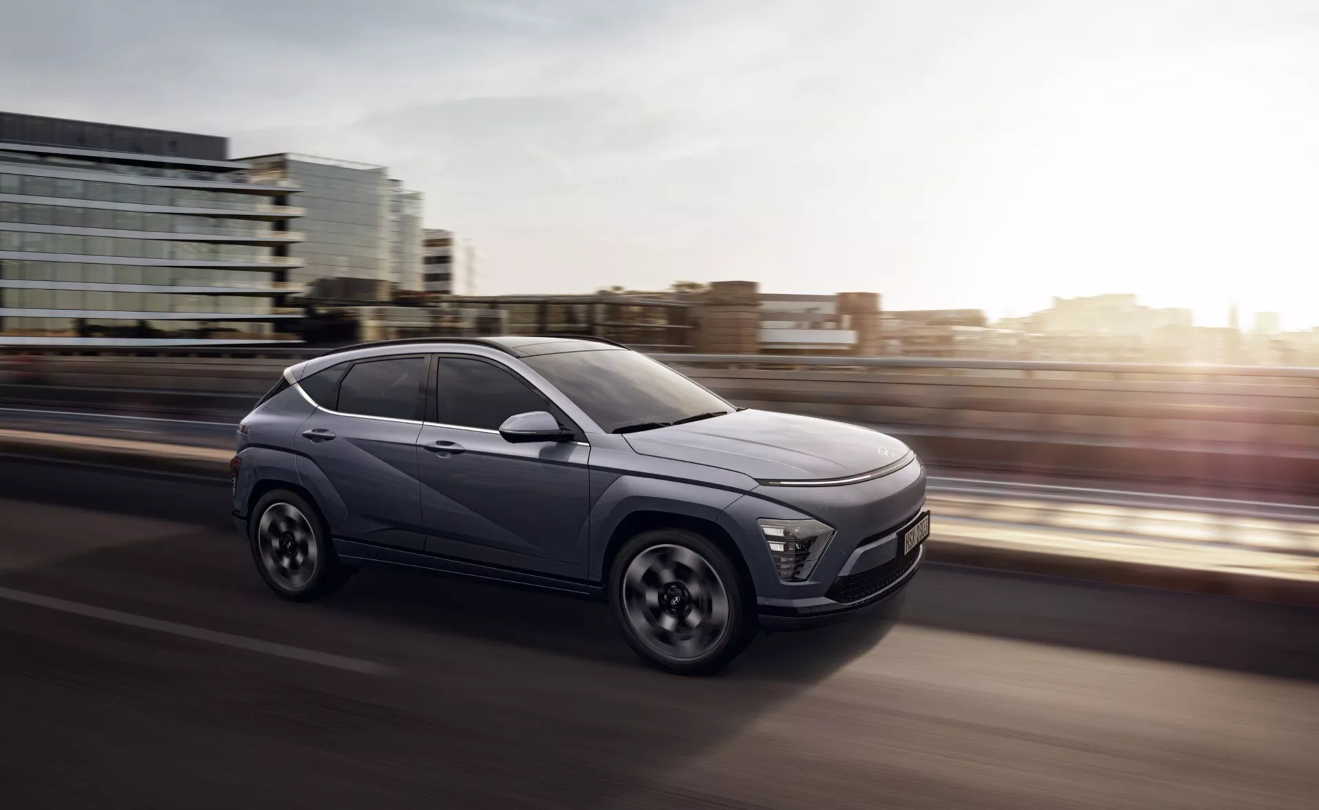 Hyundai Kona drugiej generacji 2023, czterodrzwiowy crossover SUV w ruchu, przedstawiony z dynamicznego trzykwartalnego przedniego kąta podkreślającego nowoczesne elementy projektu takie jak reflektory LED, charakterystyczna przednia atrapa wskazująca na pojazdy elektryczne, felgi aluminiowe wieloszprychowe i zaawansowane systemy wspomagania kierowcy, na tle wczesnego poranka lub późnego popołudnia podkreślające prędkość i ruch do przodu.